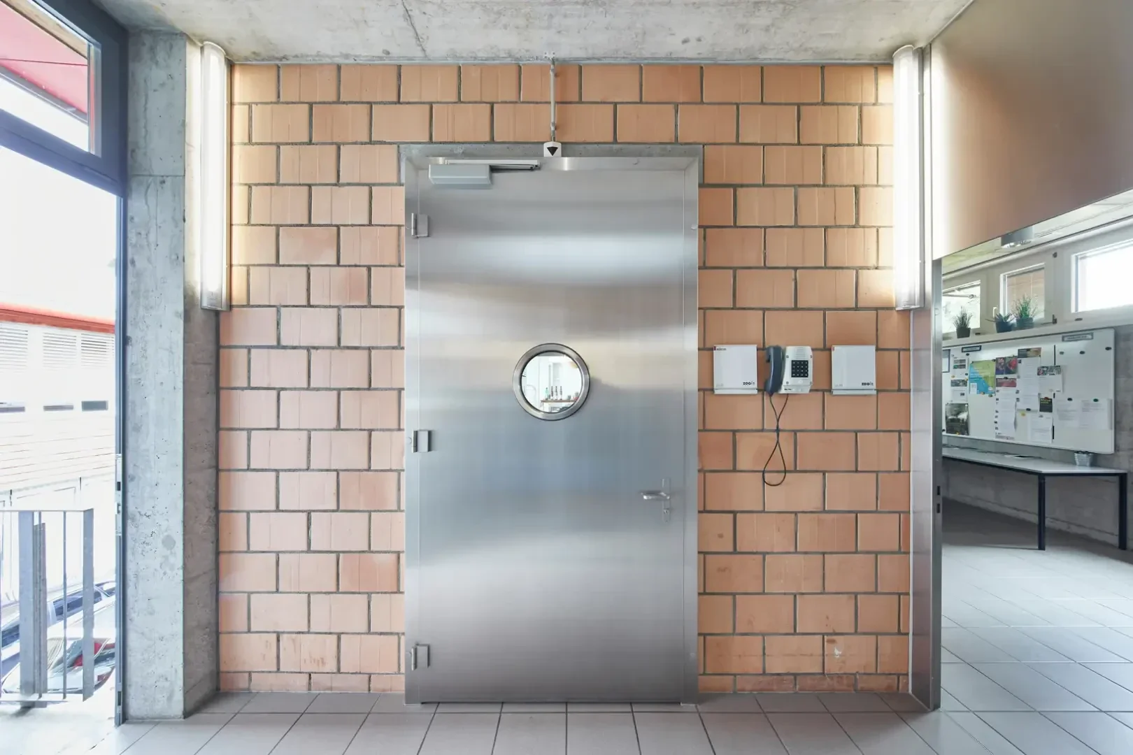 Eine große Metalltür mit einem runden Fenster ist in eine Wand aus beigen Ziegeln eingelassen. Leuchtstoffröhren rahmen die Tür ein, und an der Wand daneben sind Schalttafeln angebracht. Auf der rechten Seite ist ein schwarzes Brett zu sehen.