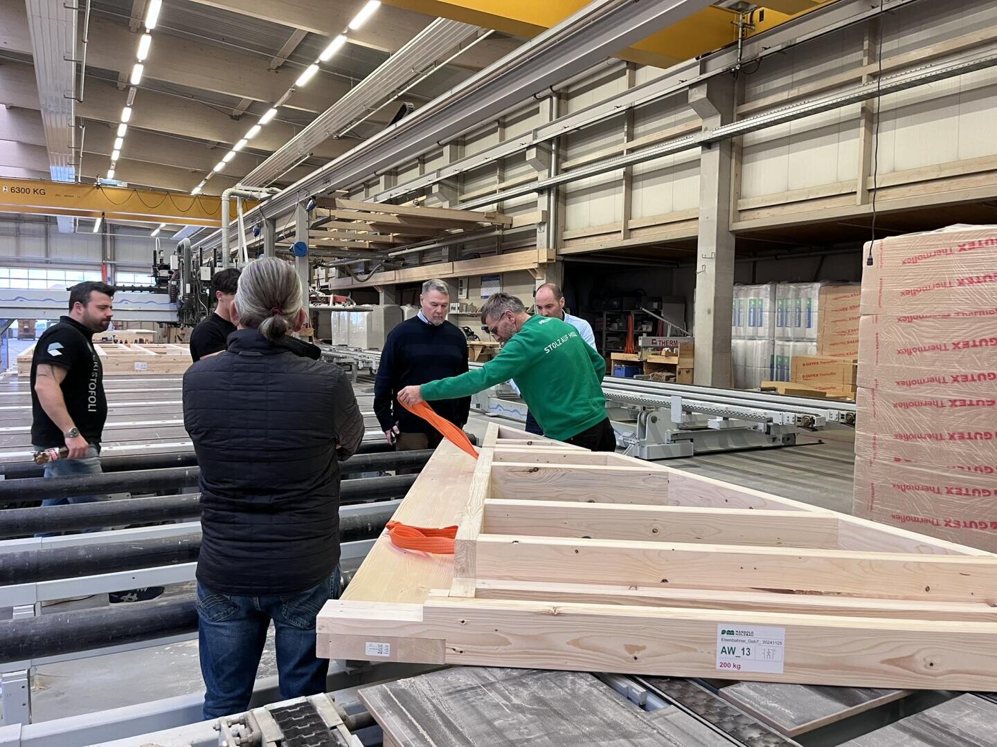 Eine Gruppe von Personen steht um einen hölzernen Dachstuhl in einer Fabrik, während eine Person in einer grünen Jacke mit einem orangefarbenen Gurt demonstriert. Im Hintergrund sind Stapel von Materialien und Maschinen zu sehen.