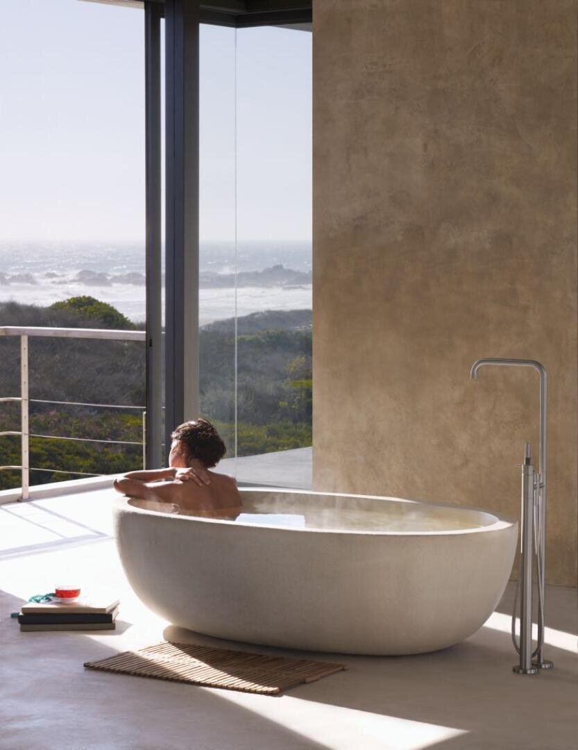 Eine Person entspannt sich in einer modernen, freistehenden Badewanne an einem großen Fenster mit Blick auf das Meer und die Küste. Sonnenlicht erfüllt den minimalistischen Raum, und ein Buch und ein Getränk liegen auf einer Matte daneben.