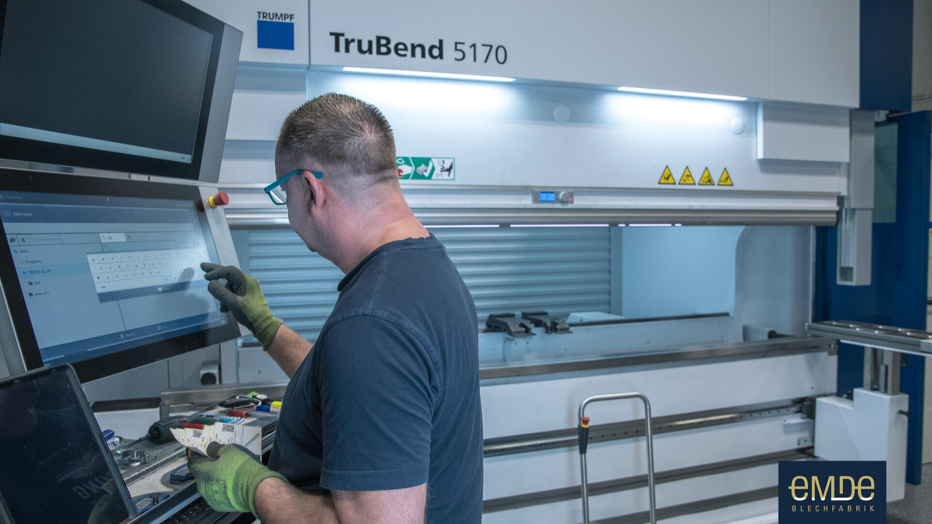 Eine Person mit Schutzhandschuhen und Brille bedient ein Touchscreen-Bedienfeld vor einer industriellen Biegemaschine TruBend 5170 in einer Fabrikumgebung. In der rechten unteren Ecke ist das eMDe-Logo zu sehen.