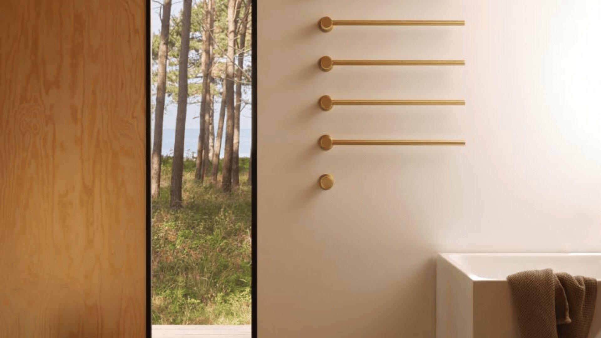 Minimalistisches Badezimmer mit vier horizontalen goldenen Handtuchhaltern an einer weißen Wand, einer weißen Badewanne mit einem braunen Handtuch und einer Tür, die den Blick auf Bäume und Grünflächen nach draußen freigibt.