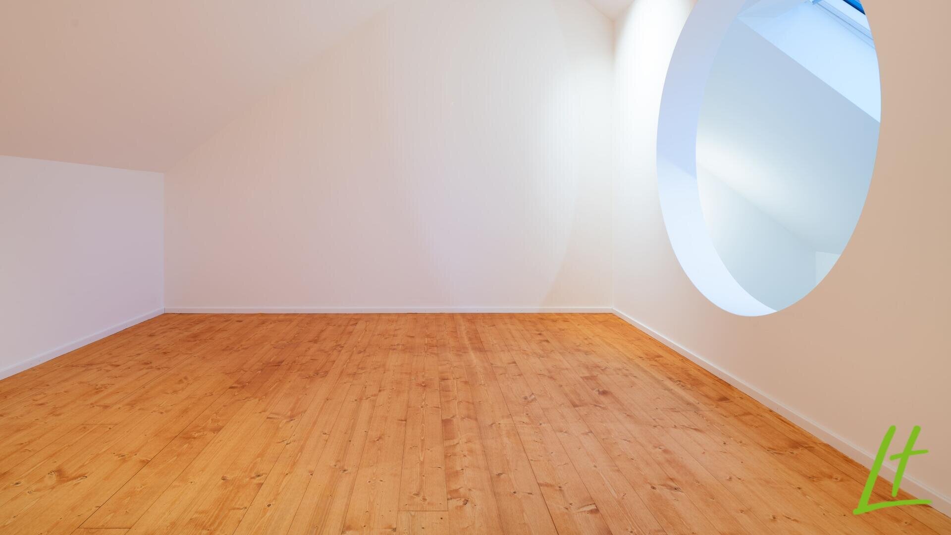 Une pièce claire et vide avec un sol en bois clair, des murs blancs et une grande fenêtre ronde qui laisse entrer la lumière naturelle. Un logo vert "lt" apparaît dans le coin inférieur droit.
