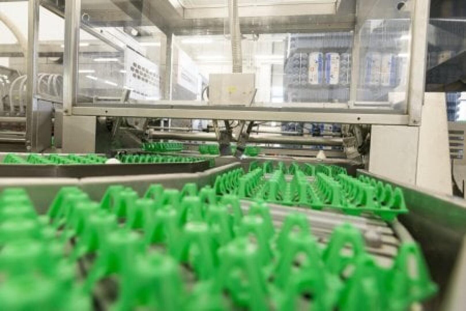 Grüne Kunststoffteile bewegen sich auf Förderbändern in einer hell erleuchteten Fabrik. Im Hintergrund sind Maschinen und transparente Schranken zu sehen, die auf einen Herstellungs- oder Verpackungsprozess hindeuten.
