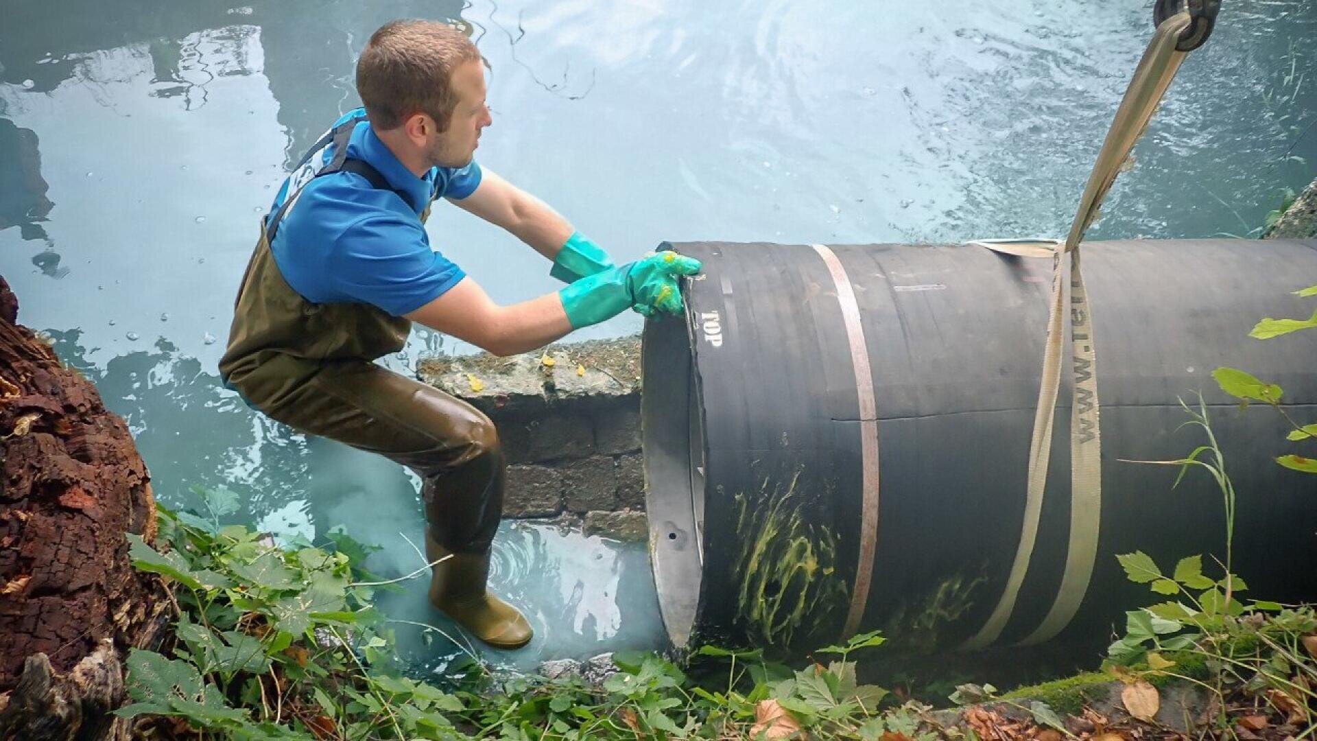 Un homme en cuissardes et gants travaille au bord d'un plan d'eau, manipulant un gros tuyau noir suspendu par une sangle. Une végétation verte entoure la zone.
