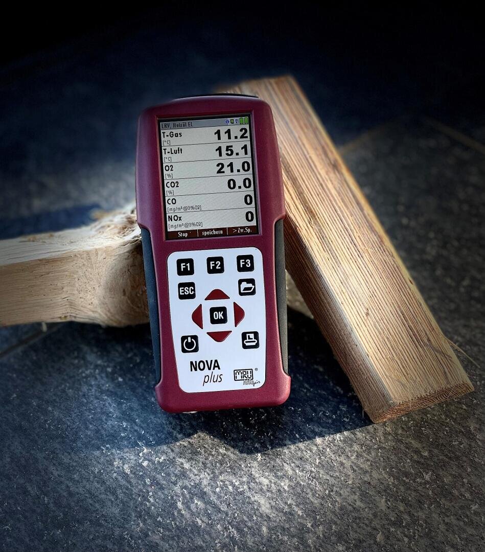 Ein tragbarer NOVA plus Gasanalysator liegt auf einer gefliesten Fläche neben zwei Holzstücken. Auf dem Bildschirm werden die Messungen verschiedener Gase angezeigt, darunter T-Gas, T-Luft, O2, CO2 und CO.