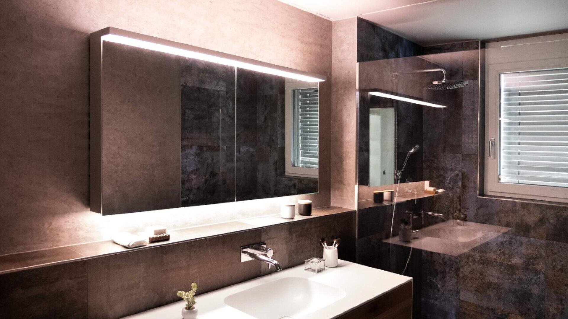Salle de bains moderne avec une grande armoire de toilette, un lavabo avec robinet, des articles de toilette sur le comptoir, une douche à l'italienne en verre et une fenêtre avec des stores qui laissent entrer la lumière naturelle. Les tons chauds et neutres et l'éclairage doux créent une atmosphère chaleureuse.