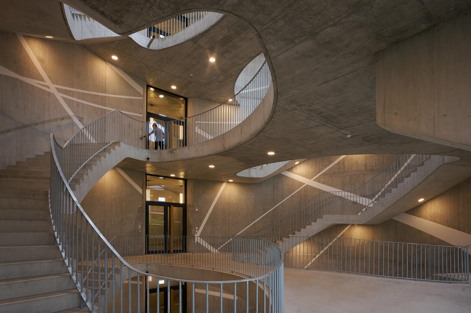 Spindeltreppen aus Beton mit Metallgeländern und geometrischen Wandmustern in einem modernen Gebäude. Warme Deckenleuchten erhellen den offenen, mehrstöckigen Raum. Eine Person steht neben einer Glastür im oberen Stockwerk.