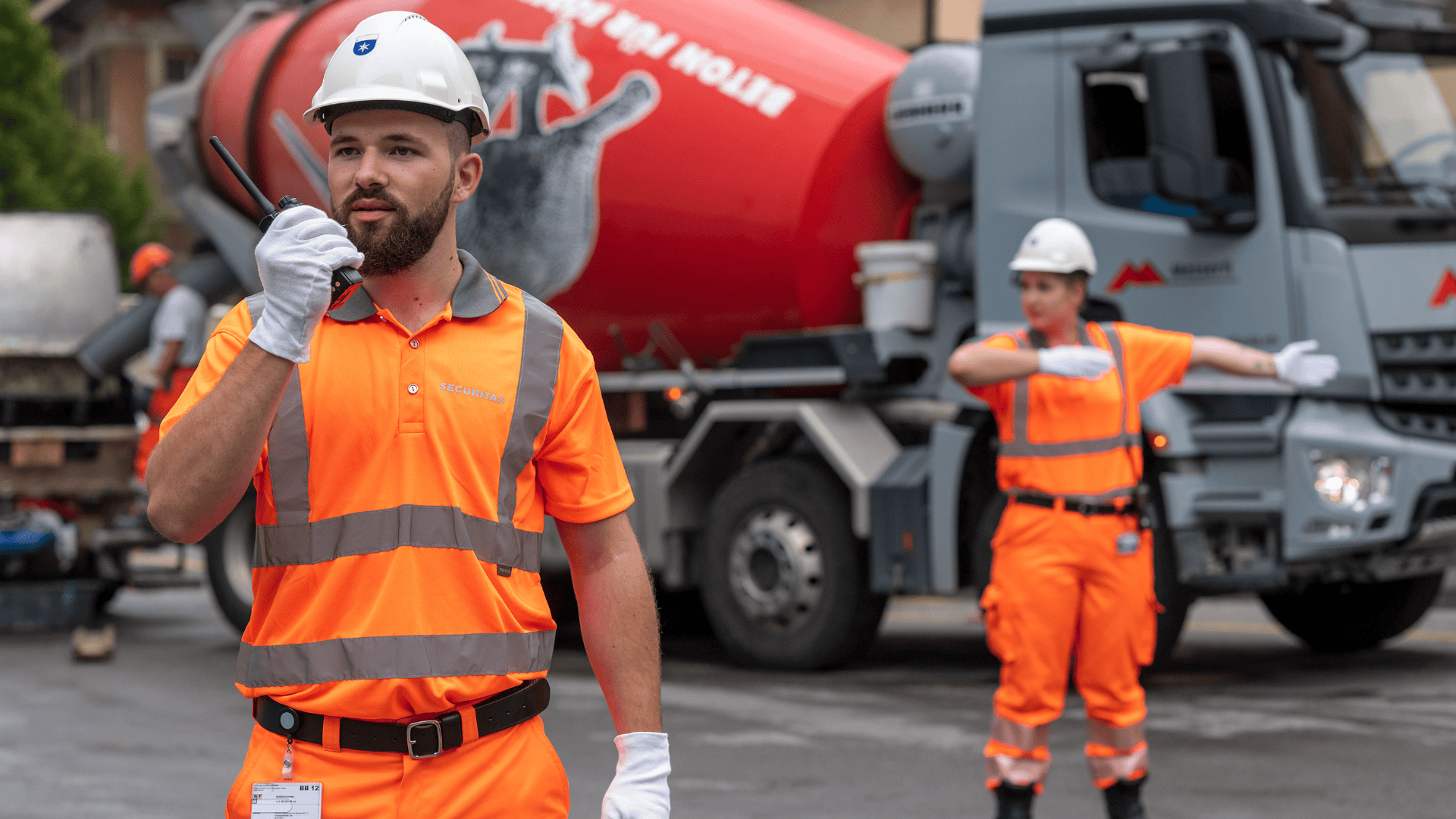 Zwei Bauarbeiter in orangefarbener Schutzkleidung und weißen Helmen leiten den Verkehr in der Nähe eines Zementlasters. Ein Arbeiter spricht in ein Walkie-Talkie, während der andere mit einem ausgestreckten Arm gestikuliert.