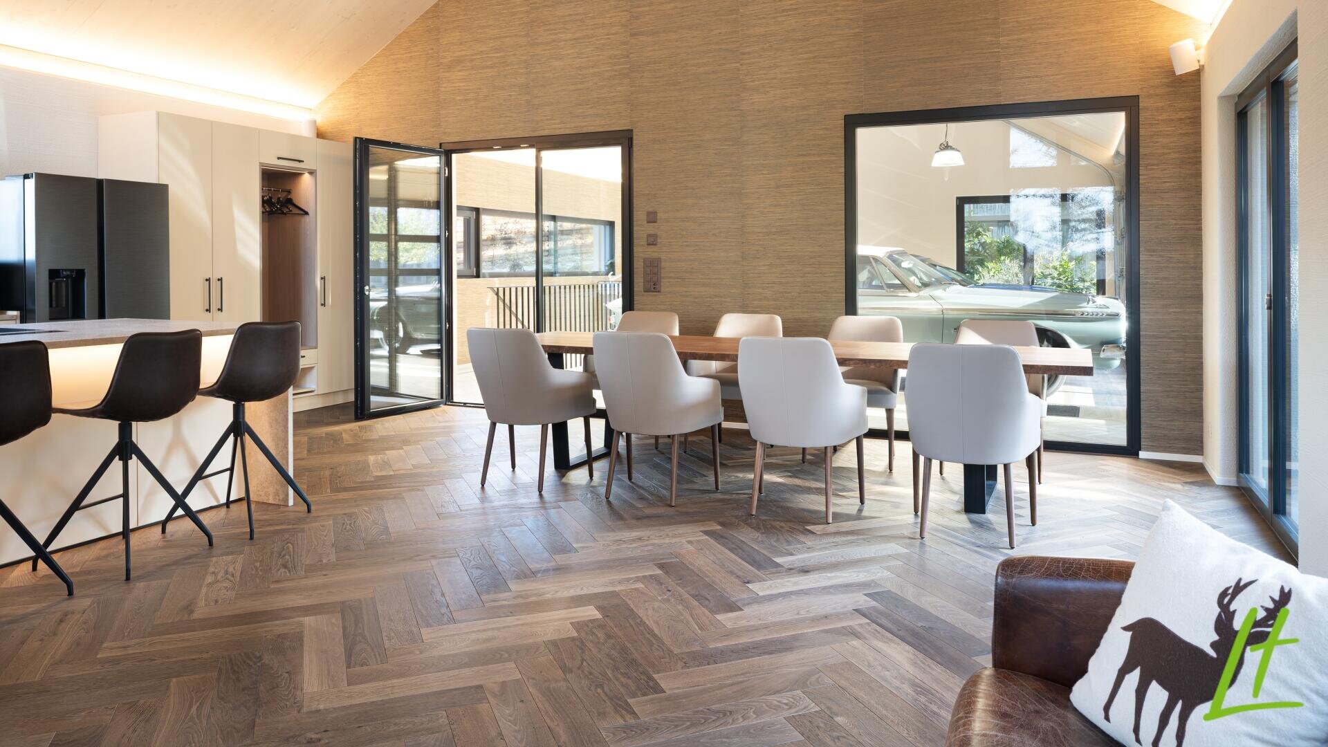 Moderner Essbereich mit einem langen Holztisch, acht weißen Stühlen, einer Küche mit schwarzen Hockern, großen Fenstern, einem Holzfußboden im Chevron-Muster und einem braunen Sessel mit einem Kissen im Hirsch-Design.