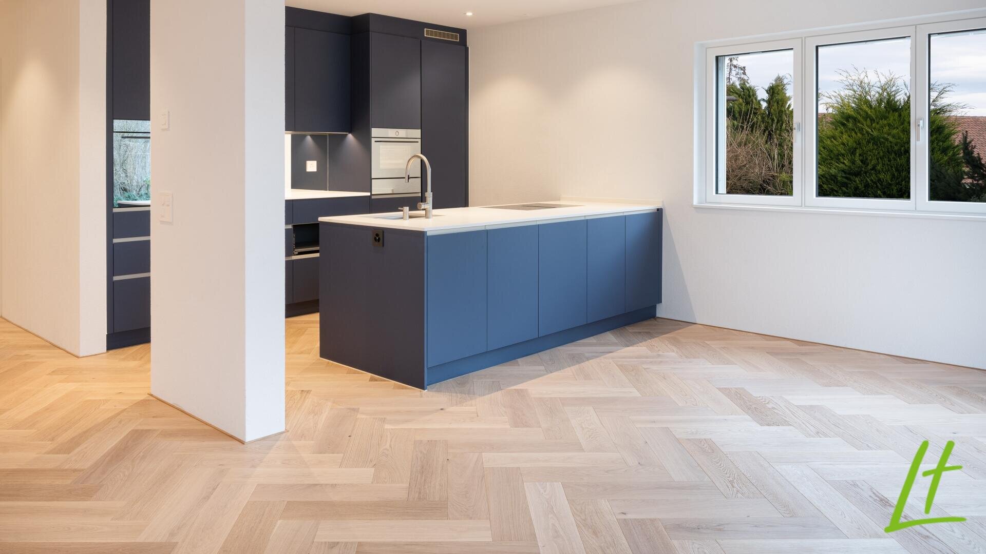 Cuisine moderne avec des armoires bleues, des plans de travail blancs, des appareils encastrés et un parquet en bois clair à chevrons. Une grande fenêtre laisse entrer la lumière naturelle et un logo vert "lt" se trouve dans le coin inférieur droit.