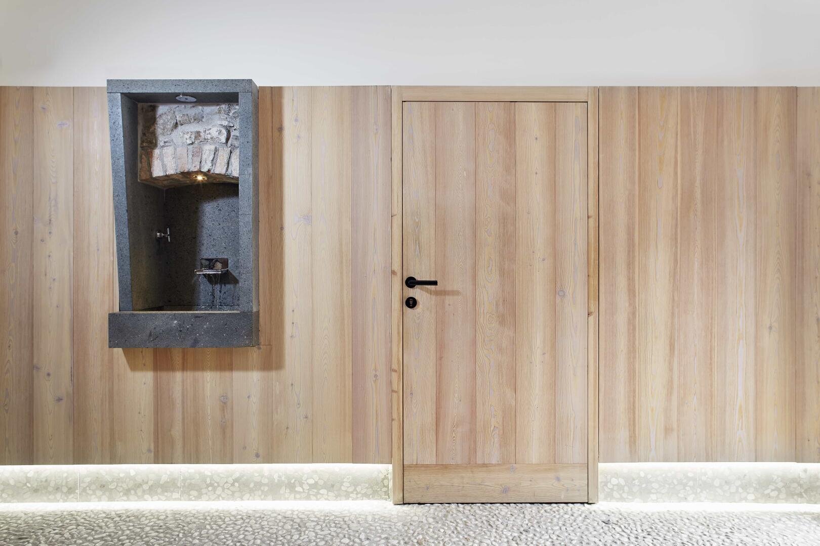 Ein modernes Interieur mit einer hellen Holzwand, einer geschlossenen Holztür mit schwarzem Griff und einem in die Wand eingelassenen Trinkbrunnen aus dunklem Stein auf der linken Seite. Der Fußboden ist mit Kieselsteinen ausgelegt, und es gibt eine sanfte Unterbeleuchtung.