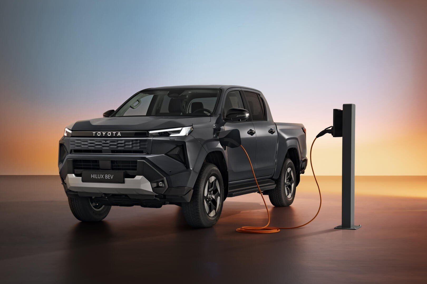 Un pick-up électrique Toyota Hilux BEV gris foncé est garé et connecté à une station de recharge, sur un fond de dégradé de couleurs dans les tons bleus, orange et jaunes.