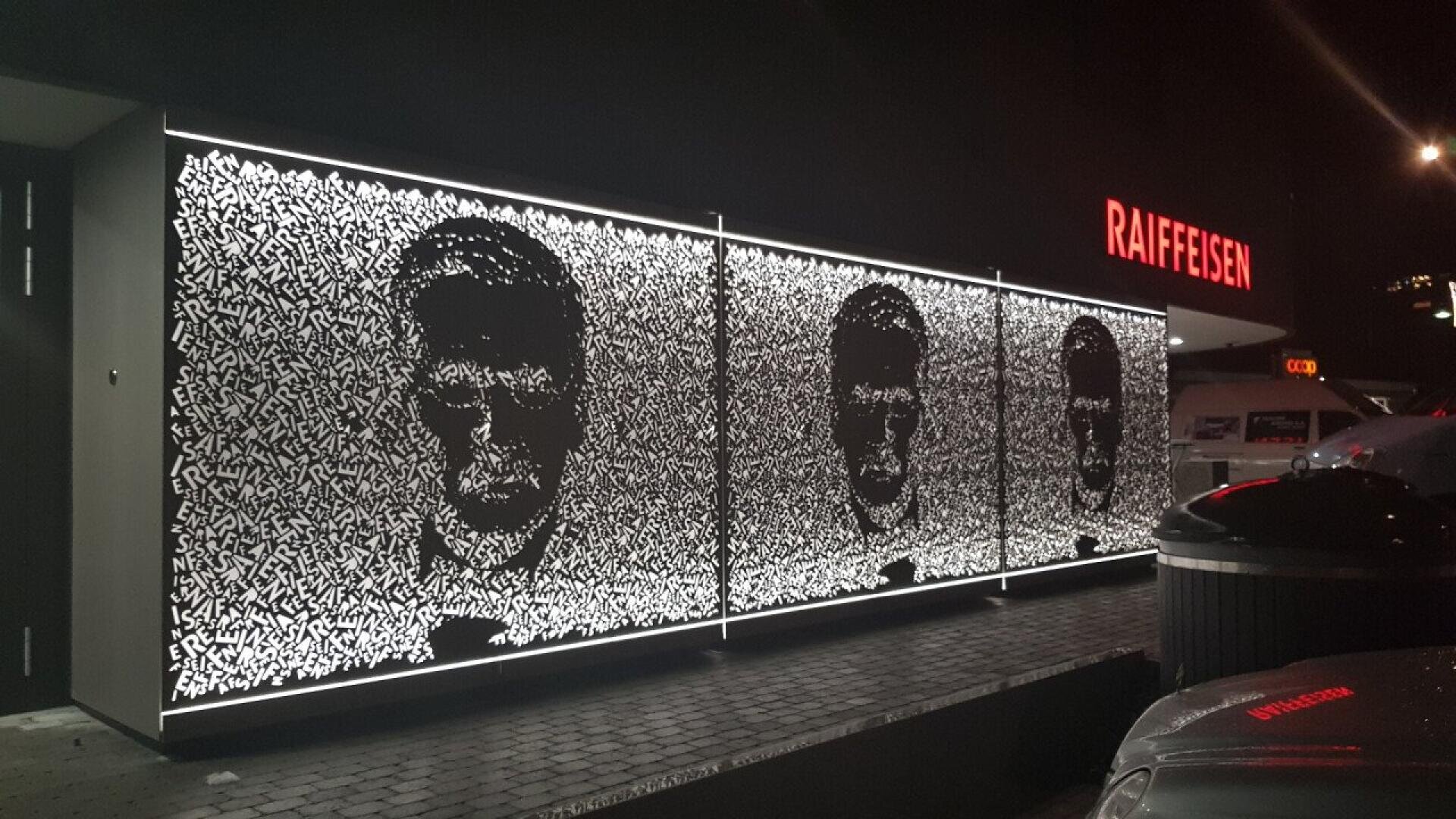 Trois panneaux lumineux présentent des portraits abstraits en noir et blanc formés par des motifs tourbillonnants. L'installation se trouve à l'extérieur, la nuit, près d'un bâtiment où se trouve une enseigne rougeoyante de RAIFFEISEN et des voitures en stationnement.