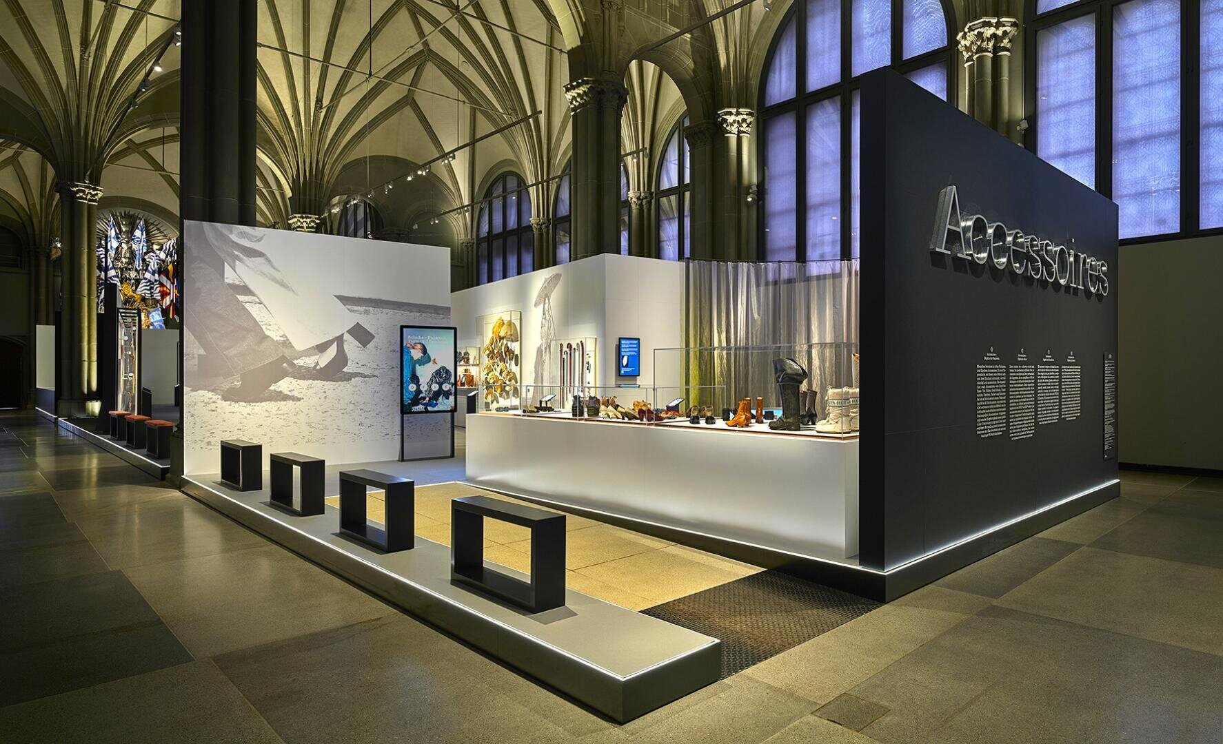 Moderne Museumsausstellung mit beleuchteten Vitrinen für Accessoires, Bänken im Vordergrund, hohen Gewölbedecken und einer großen Wand mit dem Text "Accessoires". Der Raum ist mit verschiedenen Modeartikeln und digitalen Bildschirmen ausgestattet.