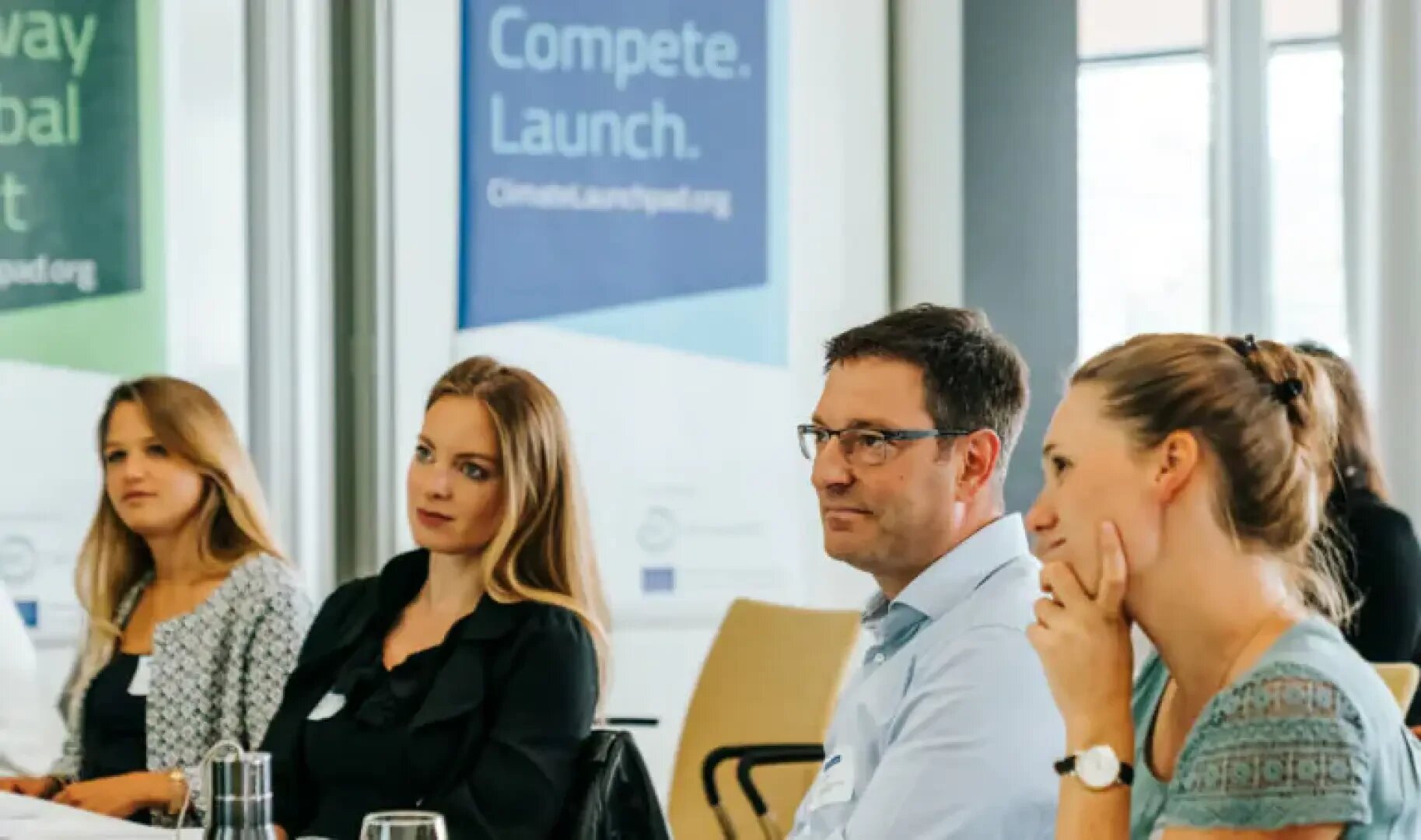 Vier Personen sitzen an einem Konferenztisch und hören aufmerksam zu. Hinter ihnen hängen große Transparente mit der Aufschrift Compete. Launch. und helle Fenster, die natürliches Licht hereinlassen.