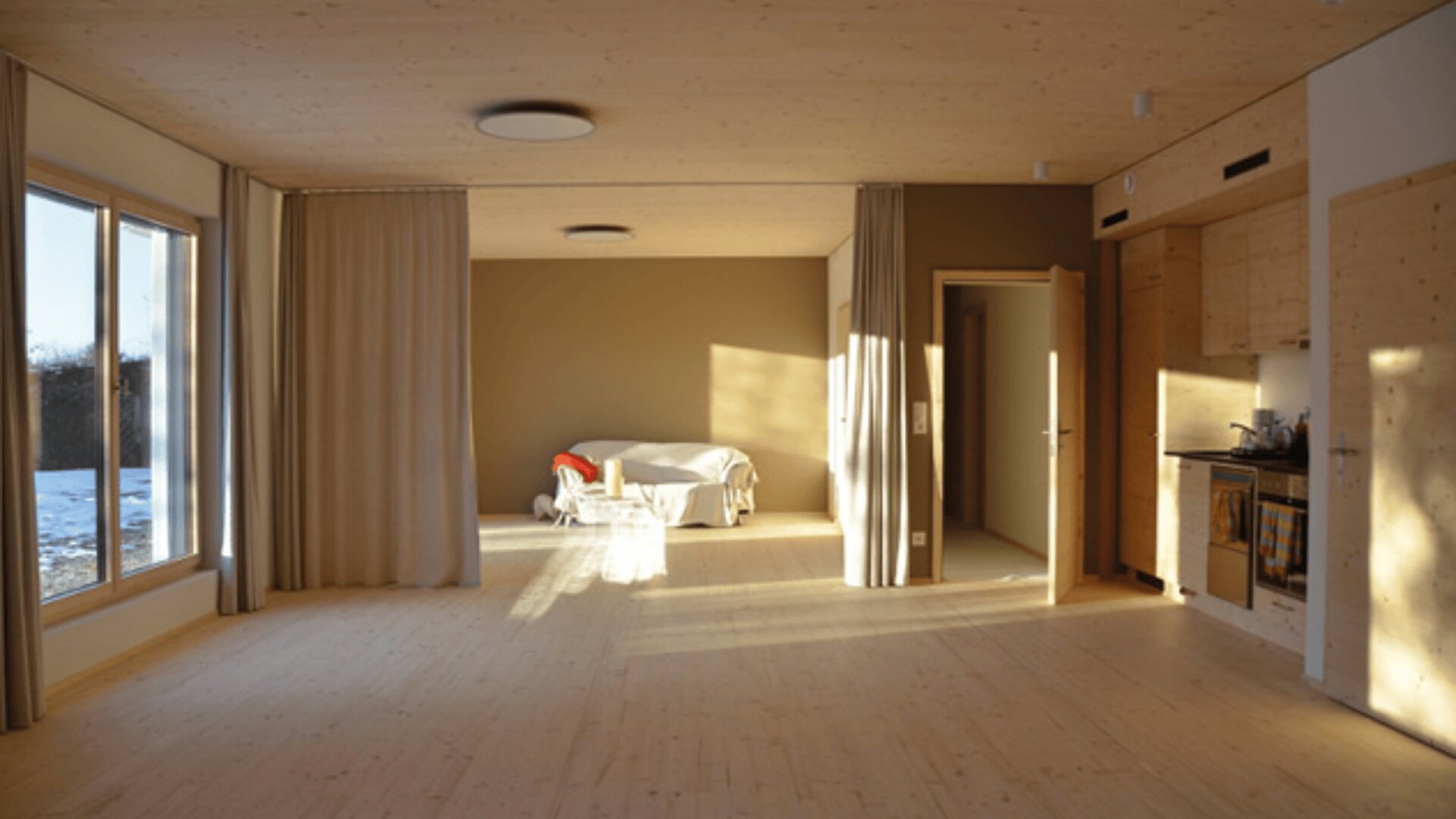 Un studio minimaliste avec des sols et des murs en bois clair, de grandes fenêtres, des rideaux beiges, un petit lit recouvert d'un drap blanc et une kitchenette compacte. La lumière du soleil pénètre à flots et projette des ombres chaudes.