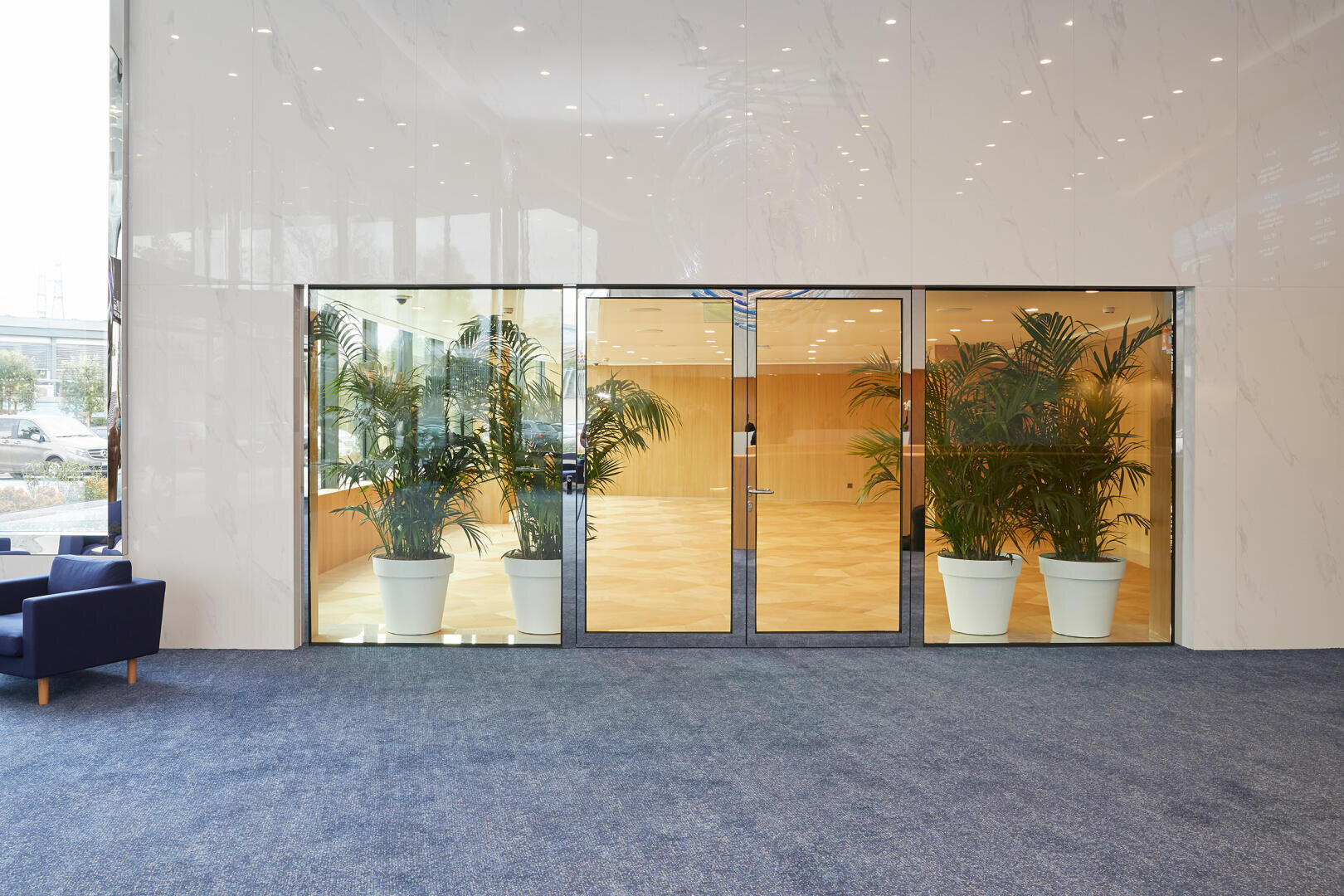 Eine moderne Lobby mit gläsernen Doppeltüren und großen Topfpflanzen auf beiden Seiten. Der Boden ist mit blauem Teppich ausgelegt, und links steht ein dunkelblauer Sessel. Der Innenraum hinter den Türen hat Holzböden und helle Wände.