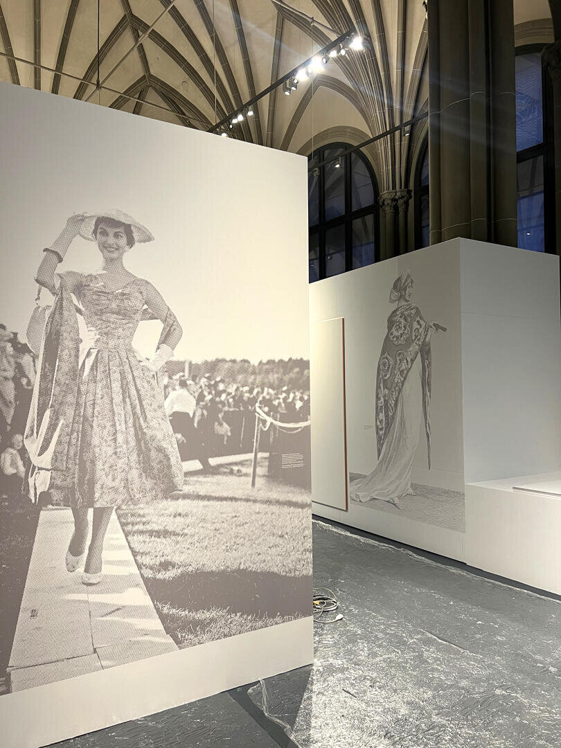 Große Schwarz-Weiß-Fotos von elegant gekleideten Frauen werden auf hohen weißen Tafeln in einem großen Ausstellungsraum mit hohen Decken, Bogenfenstern und dramatischer Beleuchtung gezeigt.