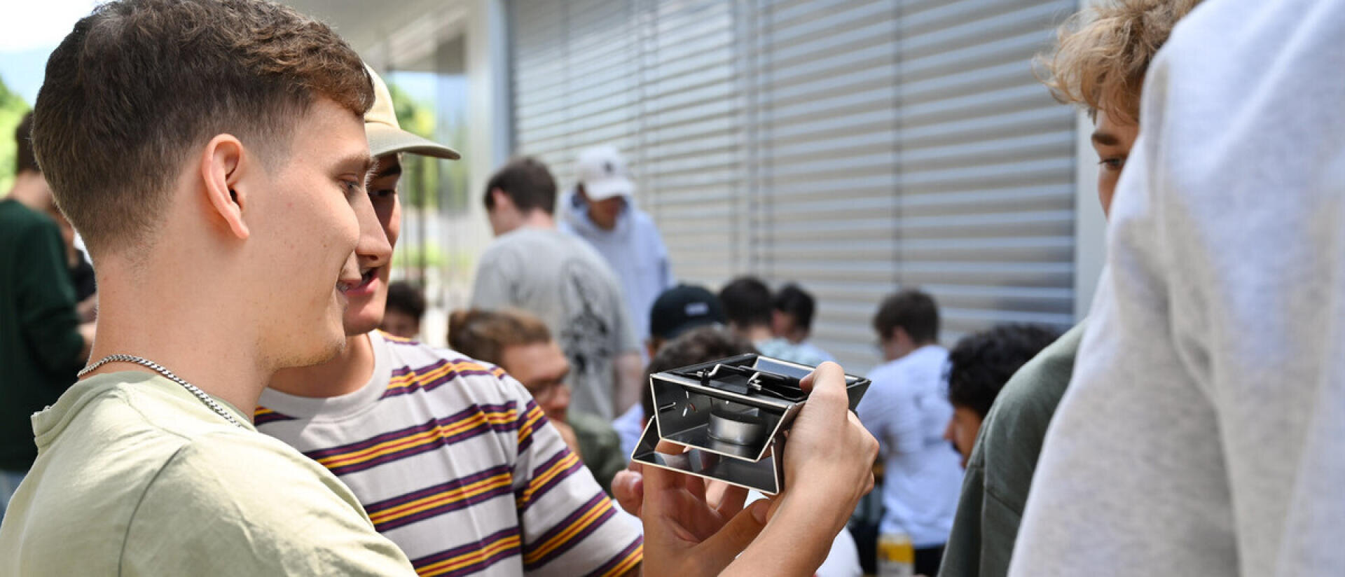 Eine Gruppe junger Leute untersucht im Freien ein kleines würfelförmiges mechanisches Objekt. Eine Person hält das Objekt hoch, während andere genau hinschauen, und im Hintergrund sind weitere Personen versammelt.