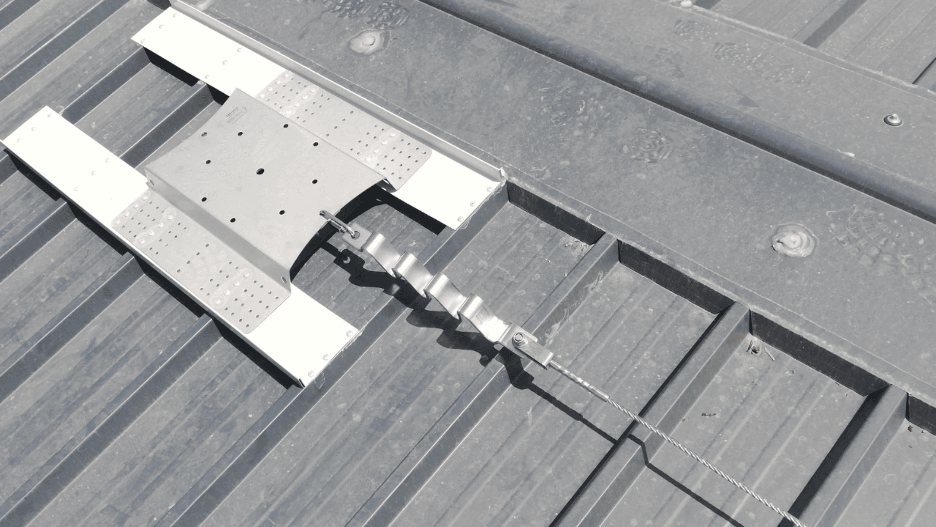 Ein Sicherheitsanker aus Metall und ein horizontales Sicherungsseil sind auf einem gerippten Metalldach installiert und dienen der Absturzsicherung bei Wartungs- oder Bauarbeiten auf dem Dach.