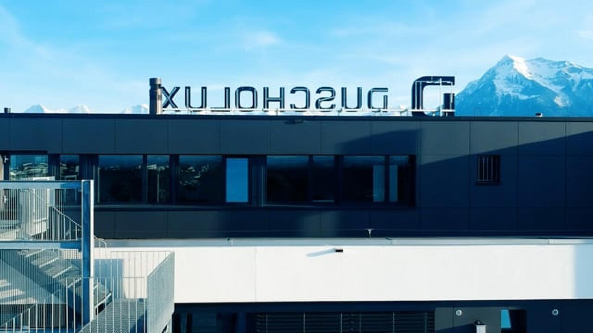 Un bâtiment moderne, gris foncé, avec de grandes fenêtres et une cage d'escalier, avec un signe LUXSCHUL inversé sur le toit ; en arrière-plan, on peut voir des montagnes enneigées et un ciel bleu.