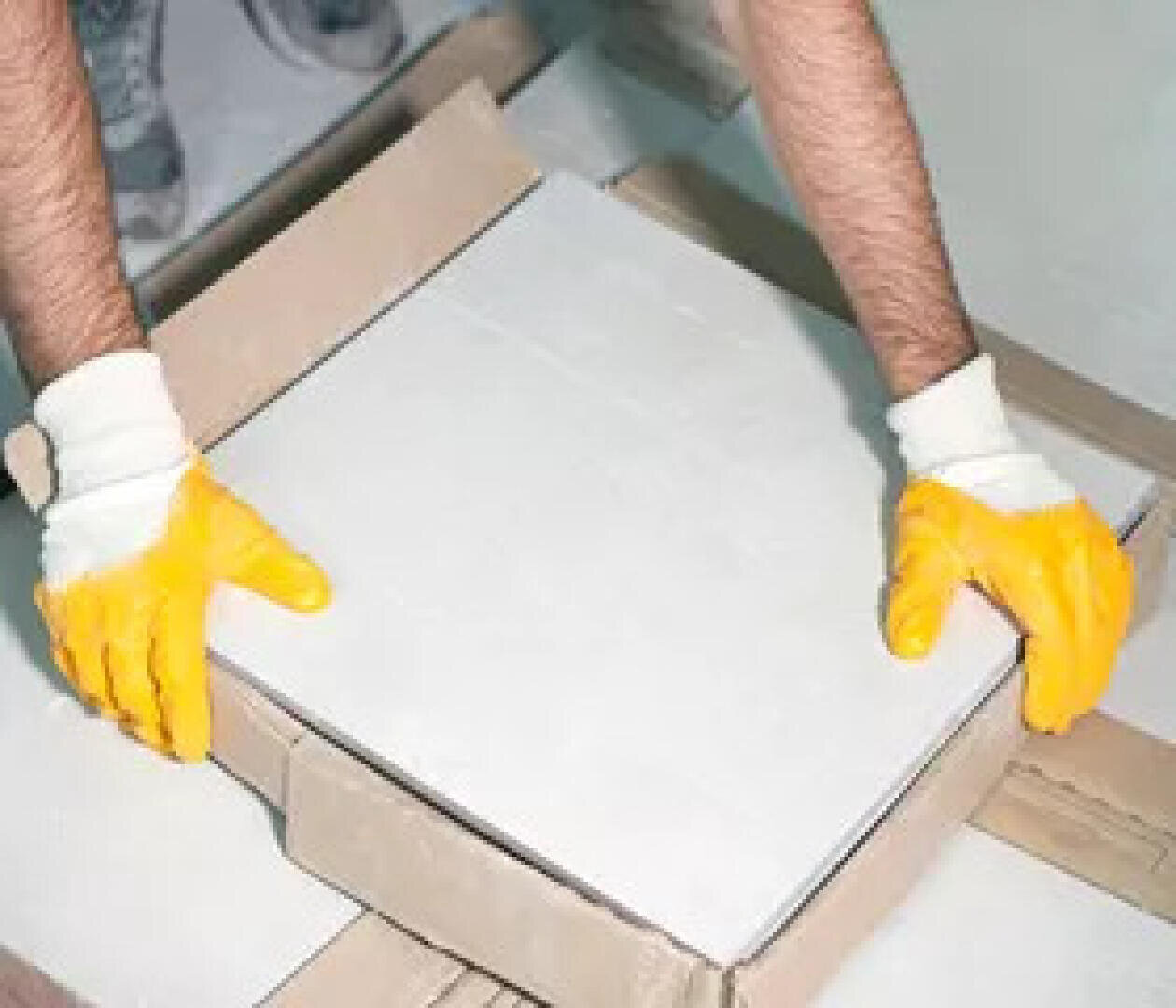 Une personne portant des gants en caoutchouc jaunes soulève ou place un grand carreau blanc dans un carton.