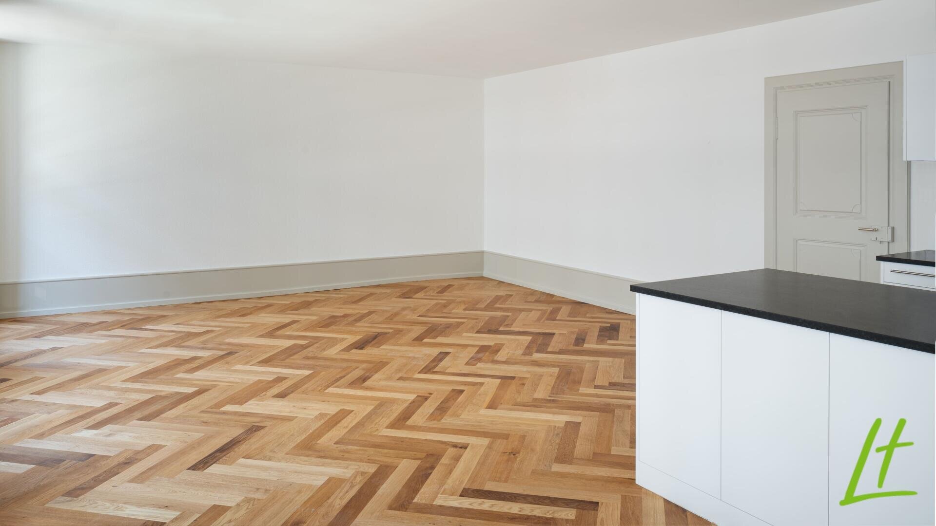 Une pièce claire et vide avec des murs blancs, un sol en bois à chevrons, une porte blanche et une kitchenette blanche avec un plan de travail noir au premier plan.