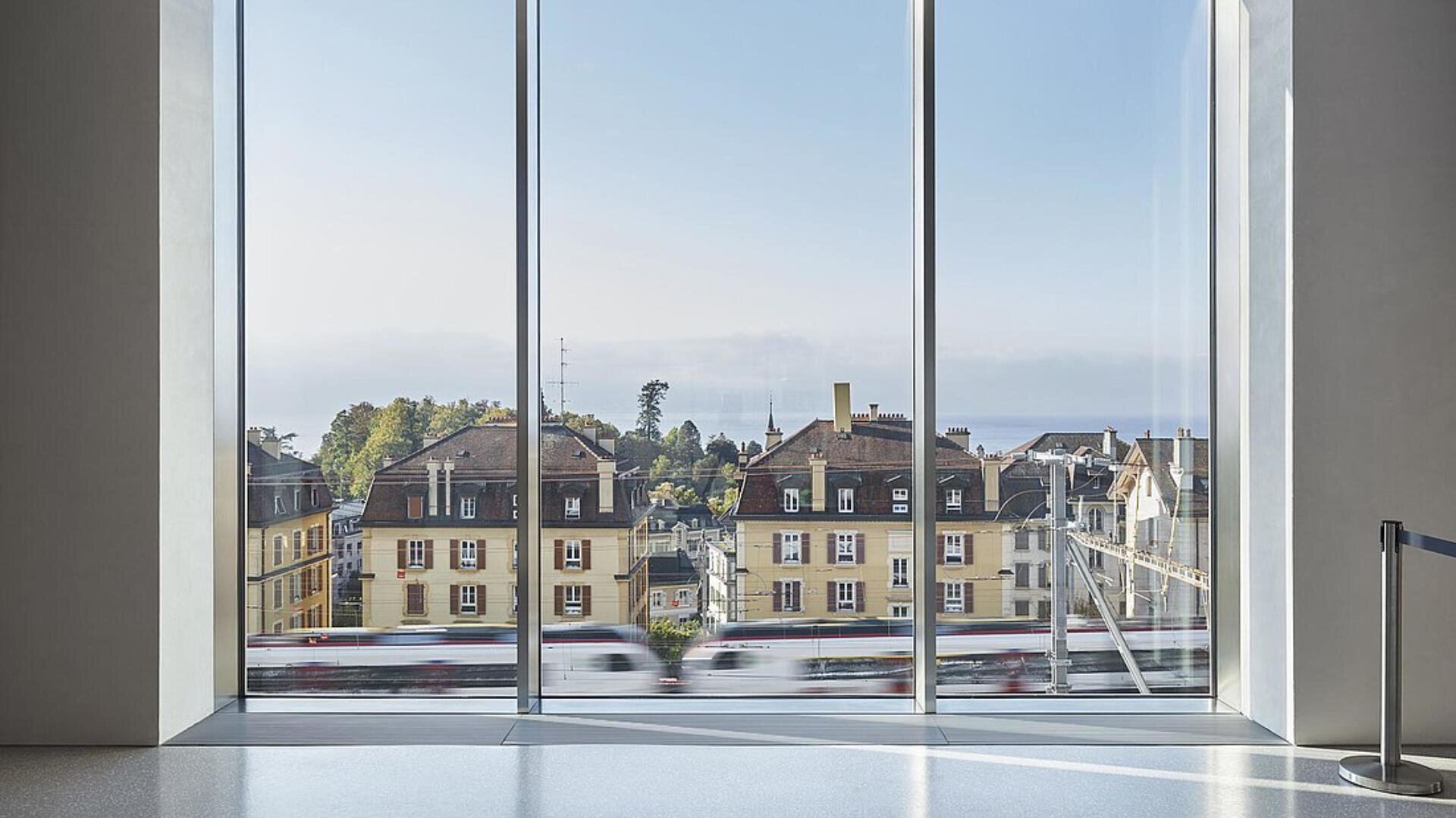Aus einem großen Fenster blickt man auf klassische europäische Gebäude mit roten Dächern und gelben Wänden. Im Vordergrund fährt ein verschwommener Zug vorbei, der Bewegung suggeriert. Der Himmel ist klar und blau, und das Sonnenlicht erfüllt den modernen Innenraum.