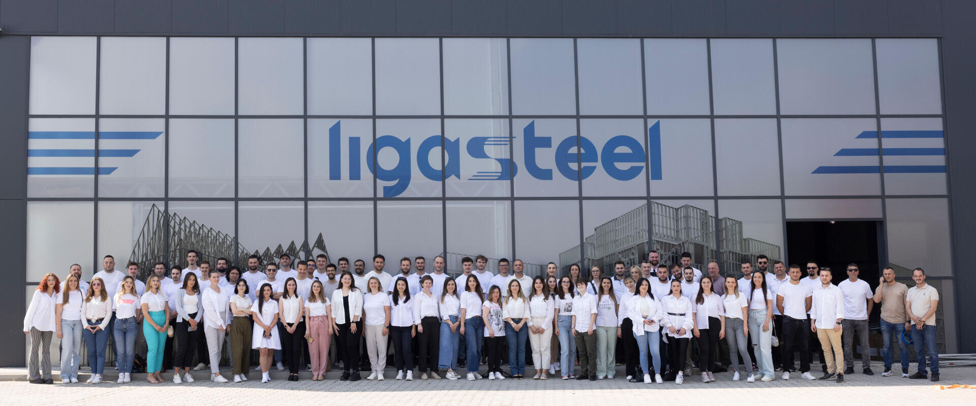 Eine große Gruppe von Menschen steht vor einem modernen Gebäude mit dem Schriftzug ligasteel an der Glasfassade. Die meisten von ihnen tragen helle Hemden und Hosen und posieren für ein Gruppenfoto auf einer gepflasterten Fläche.