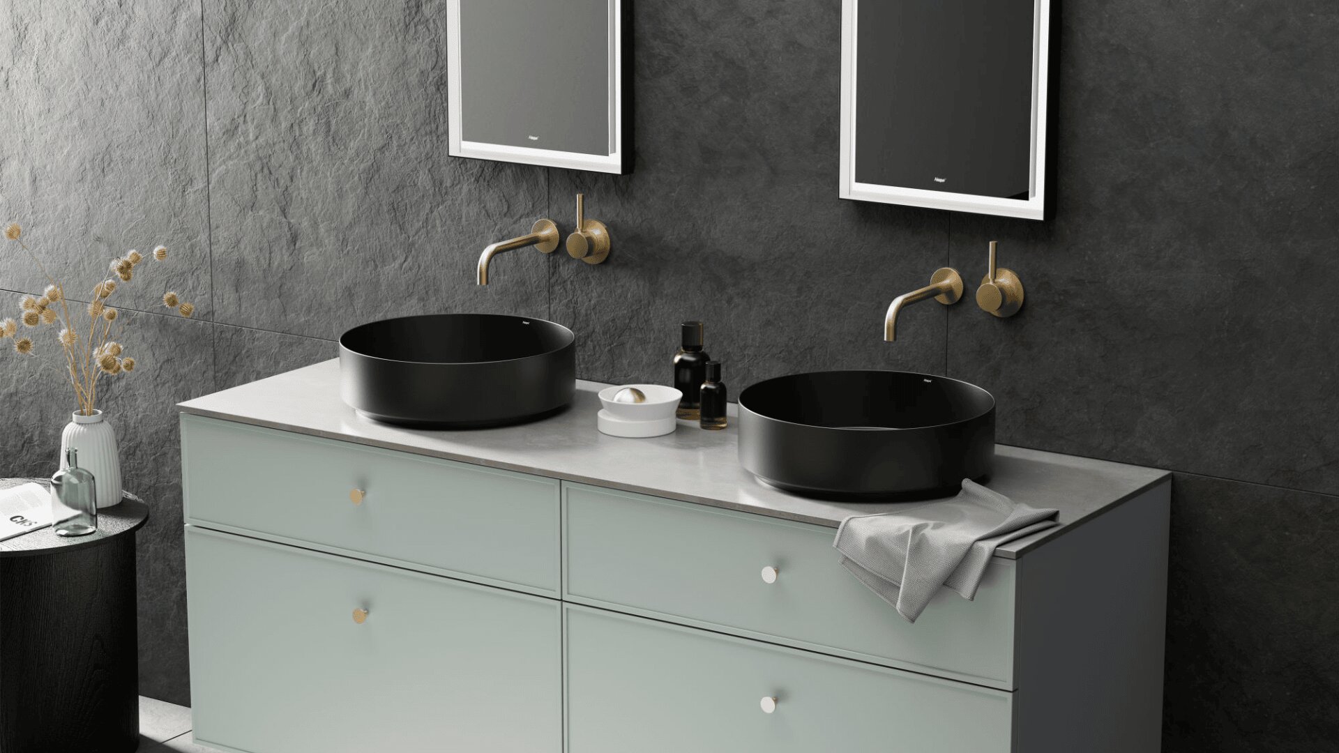 Salle de bains moderne avec deux lavabos ronds noirs, des robinets muraux en laiton, un comptoir de couleur claire, des tiroirs gris clair et deux miroirs sur un mur texturé foncé. Des bouteilles décoratives et des fleurs séchées sont également visibles.