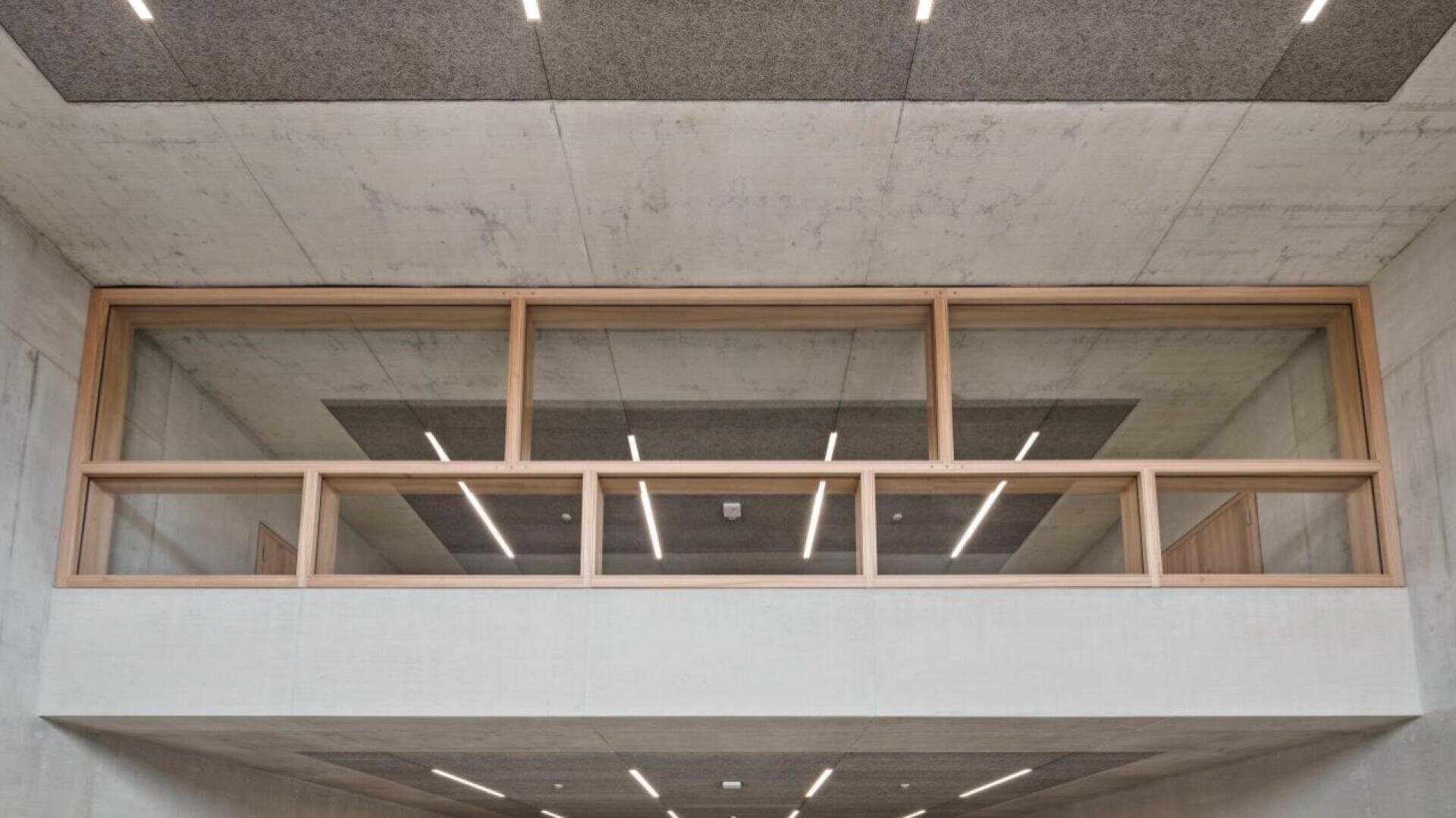 Un intérieur moderne avec des plafonds et des murs en béton et des fenêtres rectangulaires encadrées de bois donnant sur un couloir. Des plafonniers linéaires et des panneaux acoustiques soulignent le style architectural minimaliste.