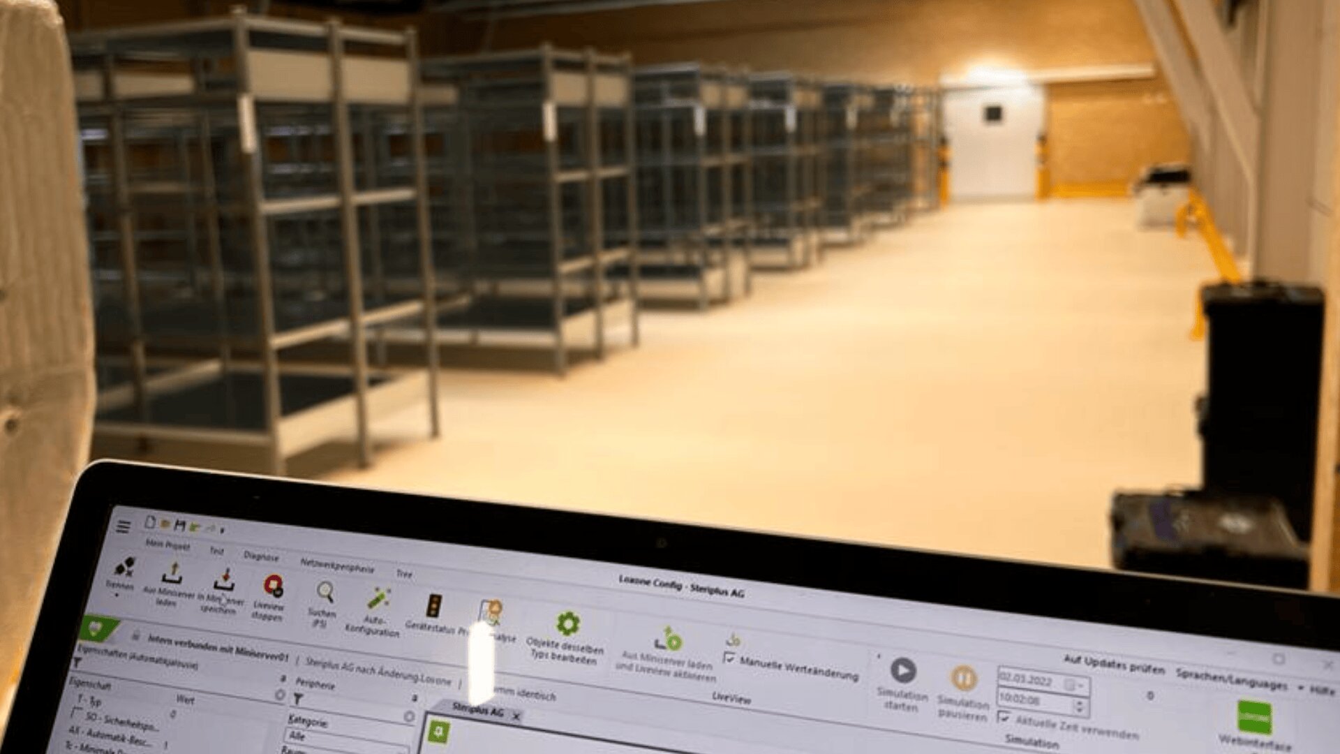Im Vordergrund ist ein Laptop-Bildschirm zu sehen, auf dem eine Logistik- oder Lagerverwaltungssoftware angezeigt wird, während im Hintergrund leere Metallregale in einem hell erleuchteten, geräumigen Lager aufgereiht sind.