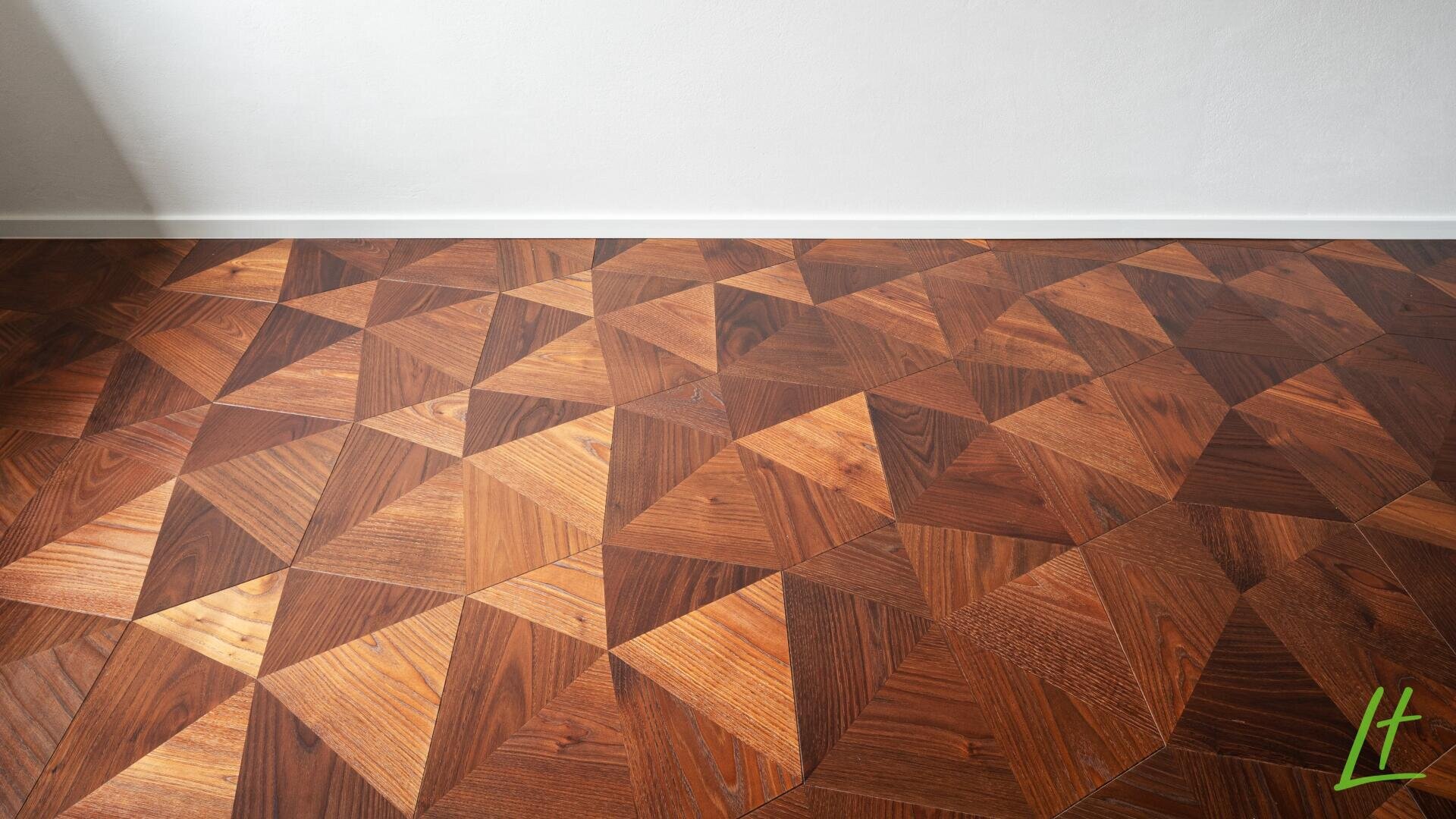 Geometrischer Holzparkettboden mit einem Muster aus ineinandergreifenden Dreiecken in verschiedenen Brauntönen vor einer schlichten weißen Wand und einer weißen Fußleiste. Ein kleines grünes Logo befindet sich in der unteren rechten Ecke.