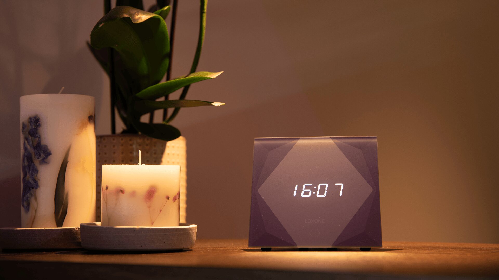 Eine Digitaluhr zeigt 16:07 auf einer hölzernen Oberfläche an, daneben stehen zwei dekorative Kerzen und eine Topfpflanze. Die weiche, warme Beleuchtung schafft eine gemütliche Atmosphäre.