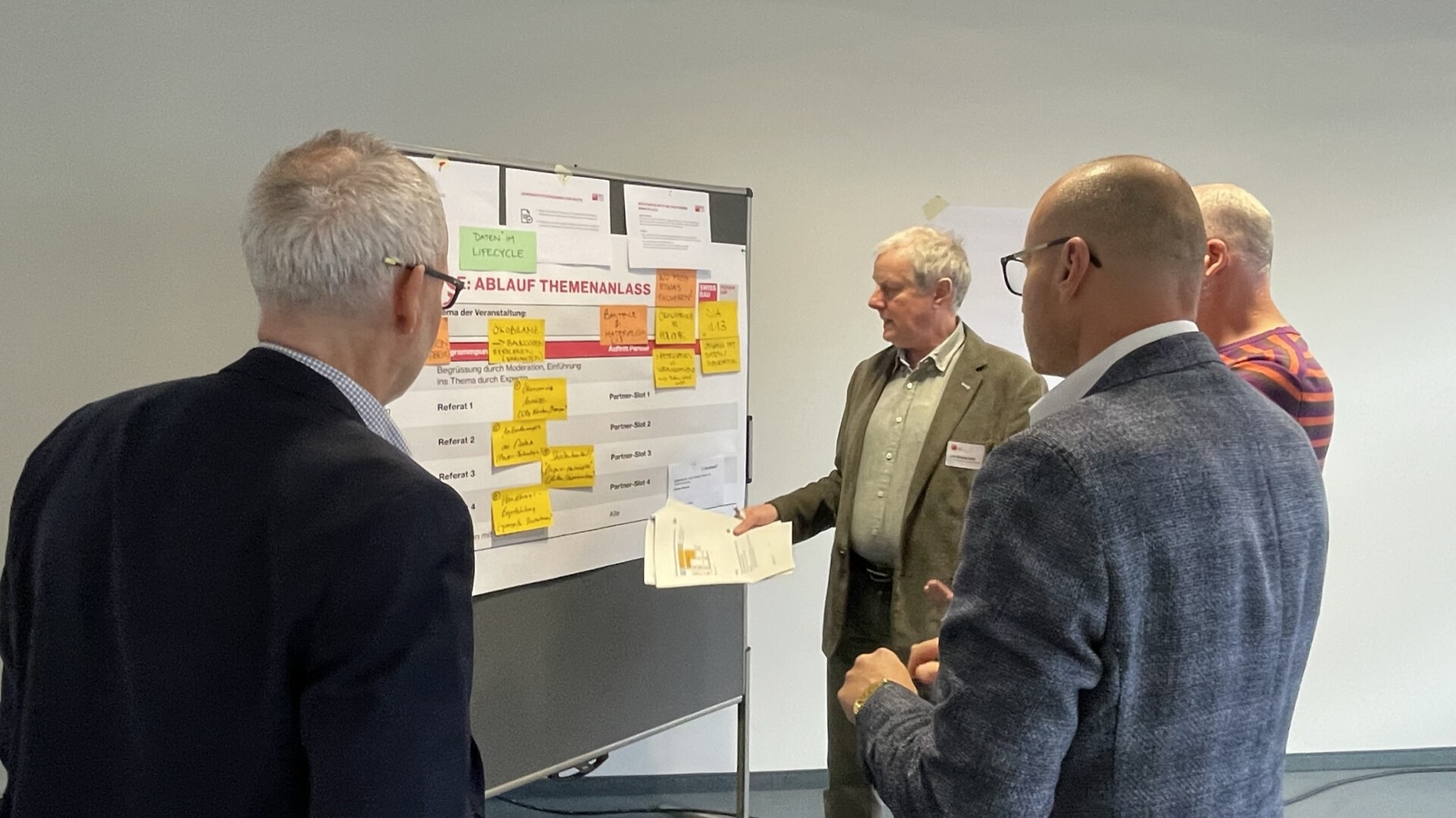 Vier Männer stehen vor einer mit Papier und Haftnotizen bedeckten Tafel, diskutieren und zeigen im Rahmen einer gemeinsamen Planungs- oder Brainstorming-Sitzung auf die Notizen.