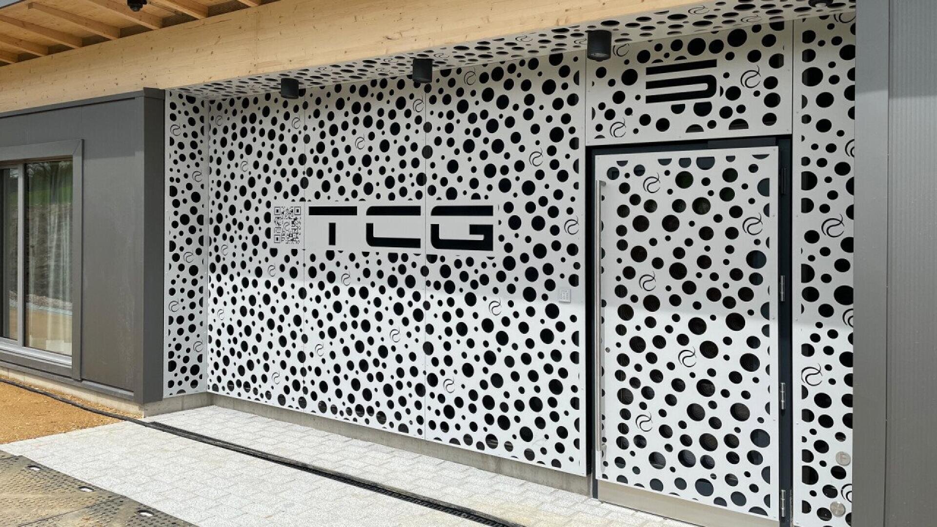 La façade du bâtiment présente un motif audacieux à pois noirs et blancs, avec une grande enseigne TCG, un code QR et un logo stylisé au-dessus d'une porte. L'extérieur est constitué d'une ossature métallique et d'une avancée de toit en bois.
