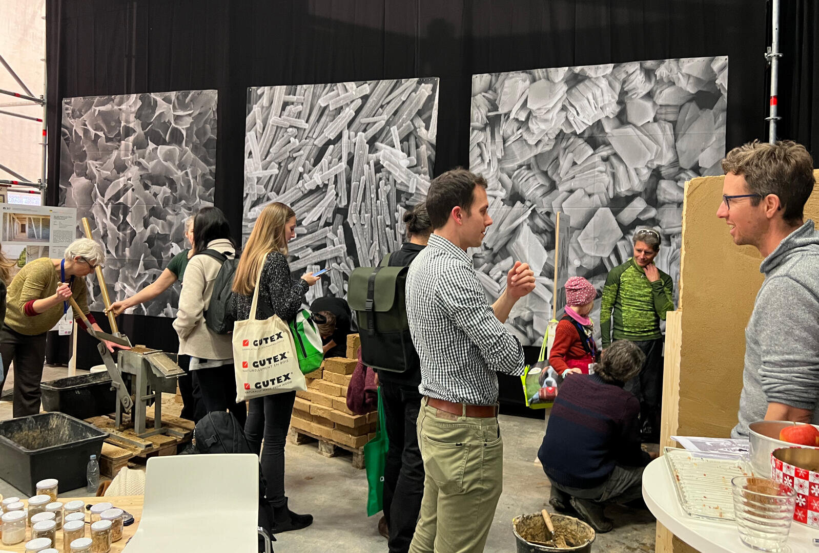 Eine Gruppe von Menschen interagiert in einer Innenausstellung mit wissenschaftlichen Displays und Materialien; große abstrakte Schwarz-Weiß-Tafeln hängen an der Wand im Hintergrund. Einige Personen unterhalten sich, während andere Gegenstände auf Tischen untersuchen.