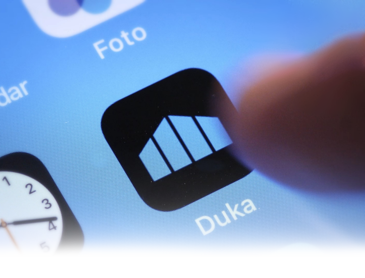 Gros plan d'un doigt qui essaie de toucher l'icône de l'application Duka sur un écran de smartphone, montrant également une partie d'une horloge et une icône d'application photo.