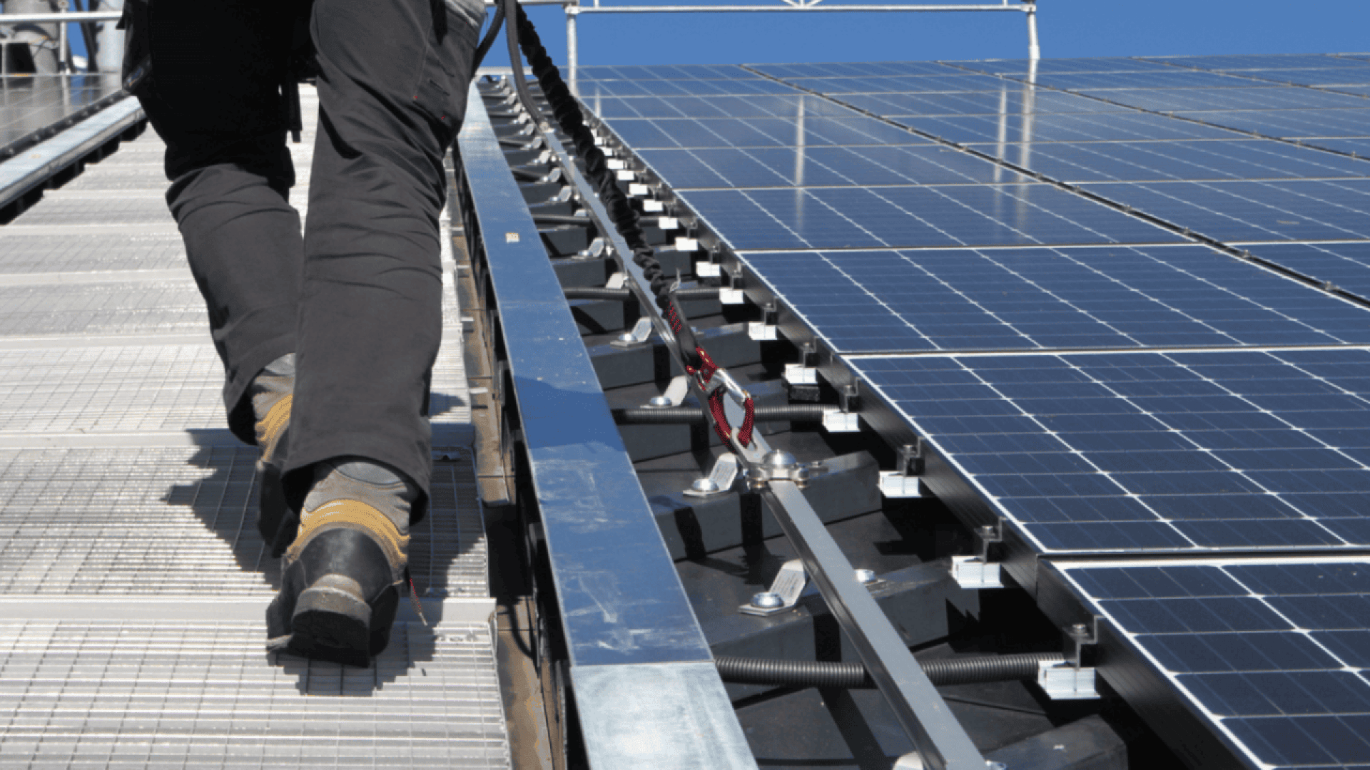 Une personne portant un pantalon sombre et des bottes de travail marche sur une passerelle métallique à côté de rangées de panneaux solaires bleus, sous un ciel bleu clair.