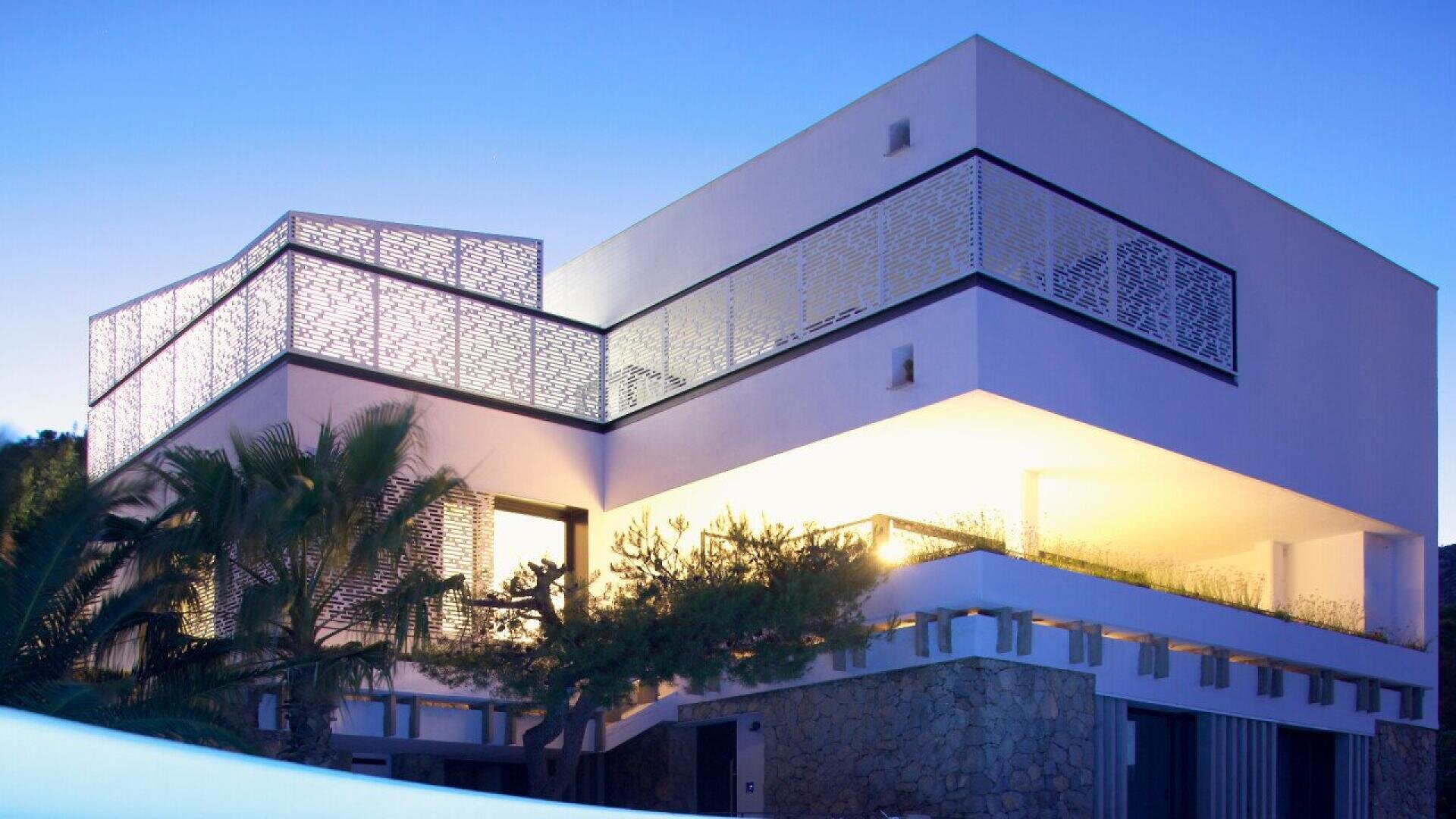 Maison moderne de deux étages aux lignes géométriques, avec de grandes fenêtres et des balustrades de balcon illuminées au crépuscule. Des palmiers et de la verdure entourent le bâtiment, et le ciel est clair et bleu.