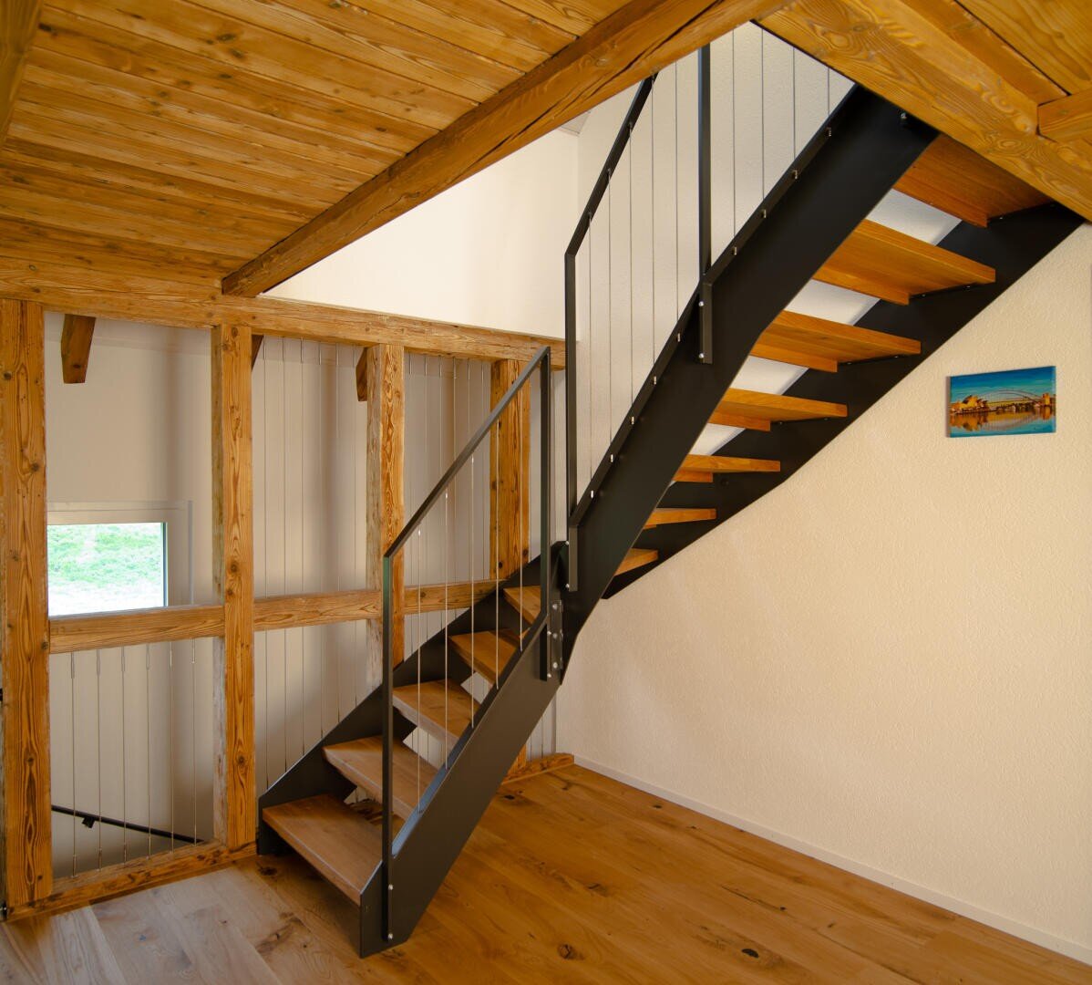 Moderne Holztreppe mit schwarzem Metallgeländer in einem Innenraum im Loft-Stil mit freiliegenden Holzbalken, hellen Holzböden, einem kleinen Fenster und einem bunten Gemälde an der cremefarbenen Wand.