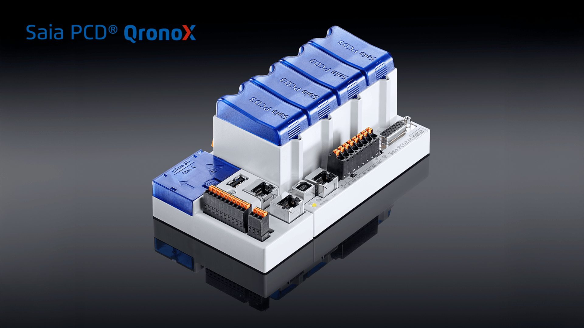 Un contrôleur d'automatisme programmable Saia PCD QronoX avec des modules bleus et blancs, différents ports et connecteurs, représentés sur une surface noire réfléchissante.