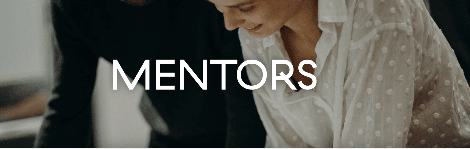 Zwei Personen arbeiten eng zusammen, eine trägt ein weißes, durchsichtiges, gepunktetes Hemd, auf dem in der Mitte in fetten weißen Buchstaben das Wort MENTORS steht.