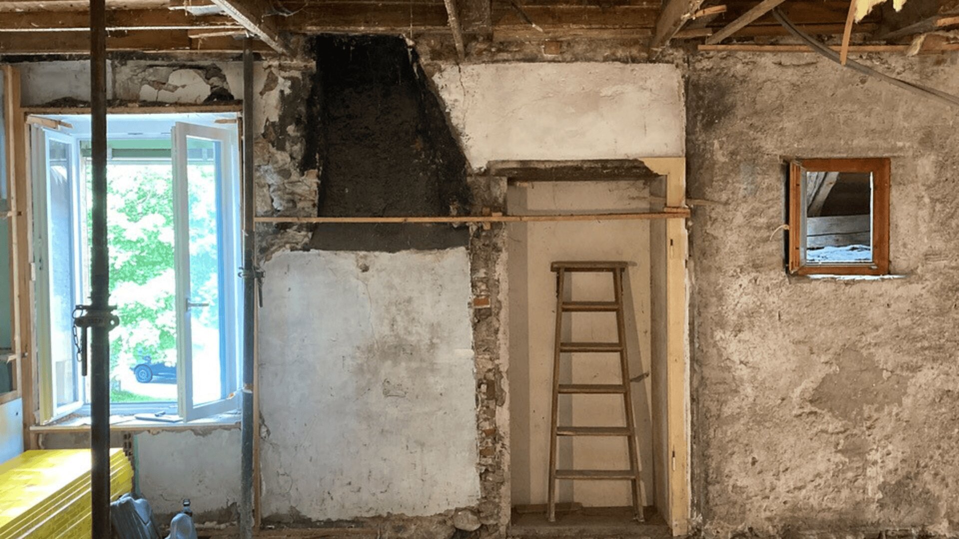 Ein renovierter Raum mit freiliegenden Backstein- und Putzwänden, einer Holzleiter an der Wand, einem offenen Fenster auf der linken Seite, durch das Sonnenlicht einfällt, und einem kleinen quadratischen Fenster auf der rechten Seite. Die Deckenbalken sind sichtbar.