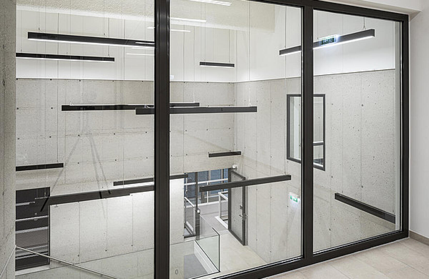 Ein modernes Interieur mit großen Glasfenstern, Betonwänden und aufgehängten rechteckigen schwarzen Leuchten. Eine Treppe und mehrere Türen sind unten in dem minimalistischen Raum im Industriestil zu sehen.