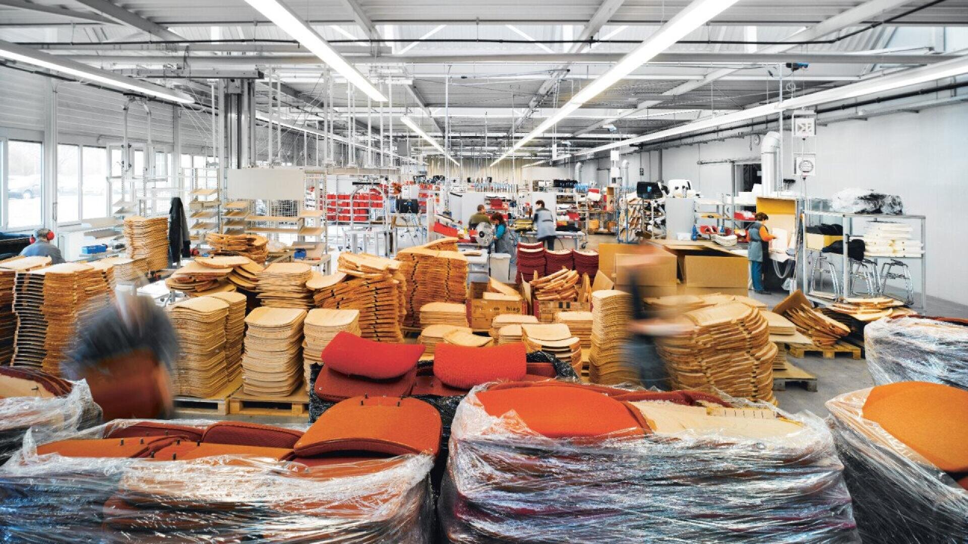 Un intérieur d'usine très éclairé avec des piles de coussins de chaise orange et beige, des travailleurs occupés, des machines et des postes de travail organisés dans un espace de travail spacieux et moderne.