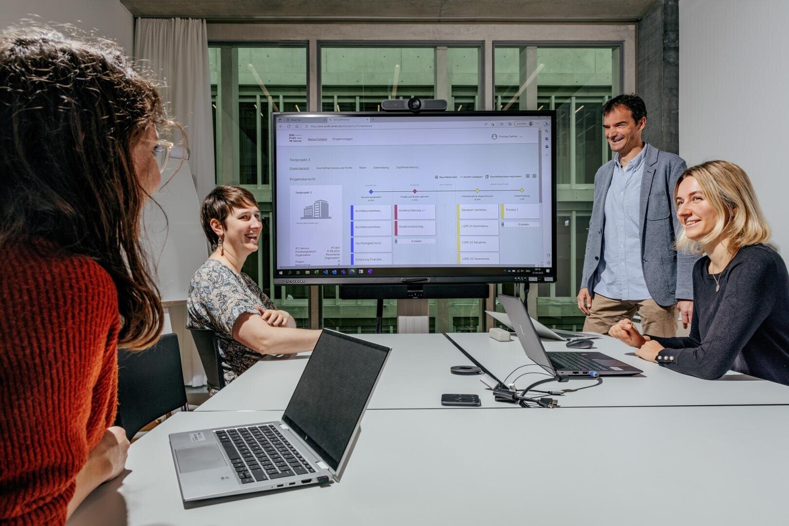 Vier Personen sitzen und stehen um einen Konferenztisch mit Laptops, lächeln und diskutieren neben einem großen Bildschirm, auf dem Diagramme und Daten in einer modernen Büroumgebung angezeigt werden.