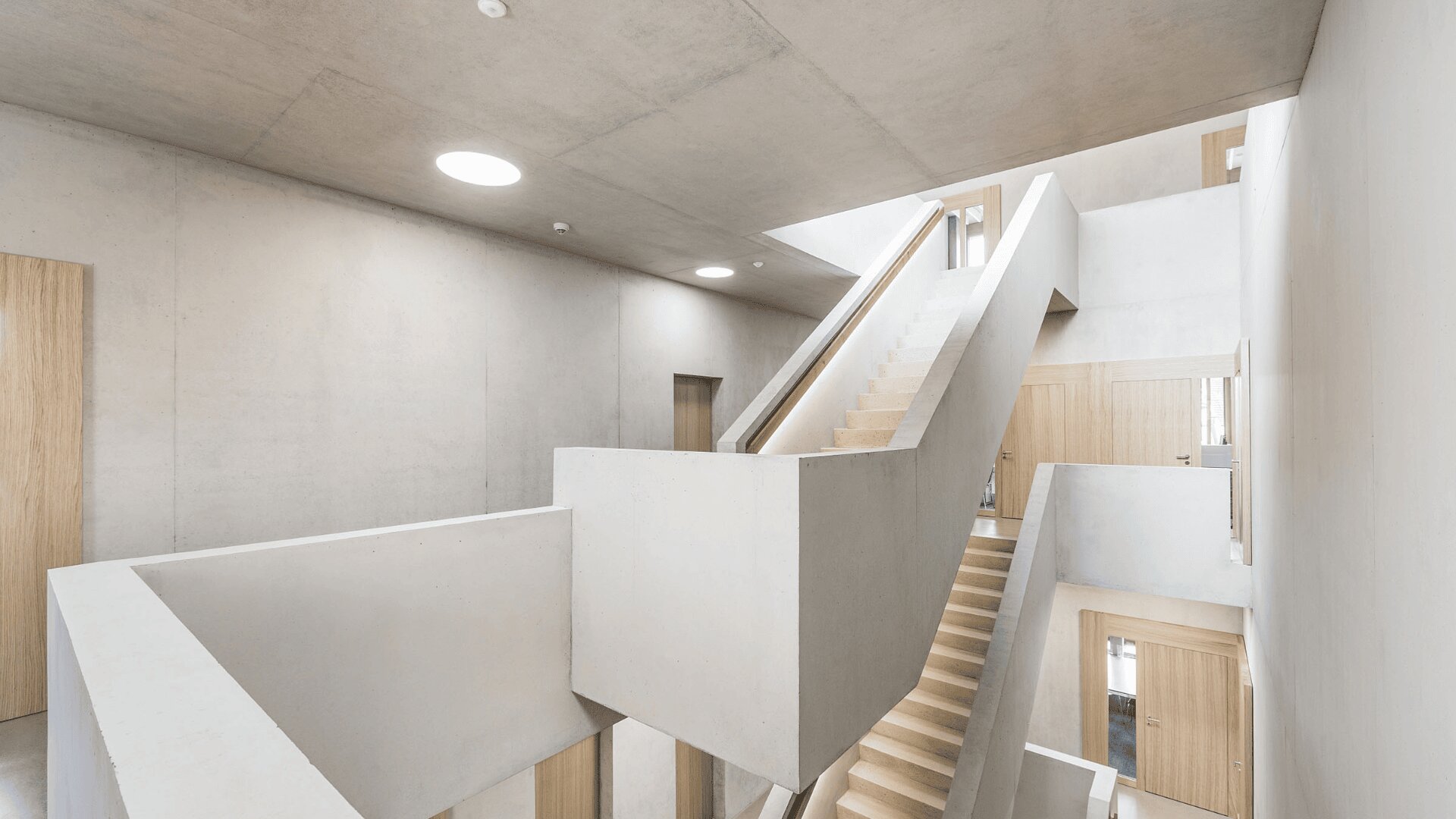 Ein modernes, minimalistisches Interieur mit weißen Betonwänden und -decken, mit mehreren Holztreppen, Holztüren und viel natürlichem Licht aus den Fenstern darüber.