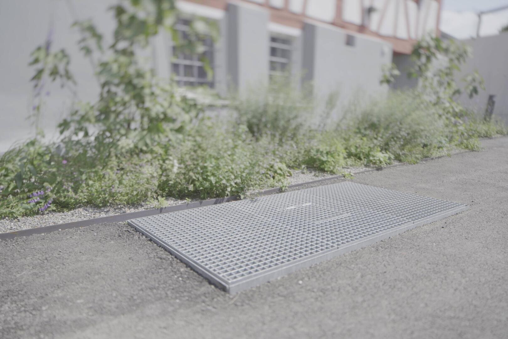 Eine Metallgitterabdeckung, die in einen gepflasterten Weg neben einem kleinen Garten mit Grünpflanzen eingelassen ist, vor einem hellen Gebäude mit Fenstern.