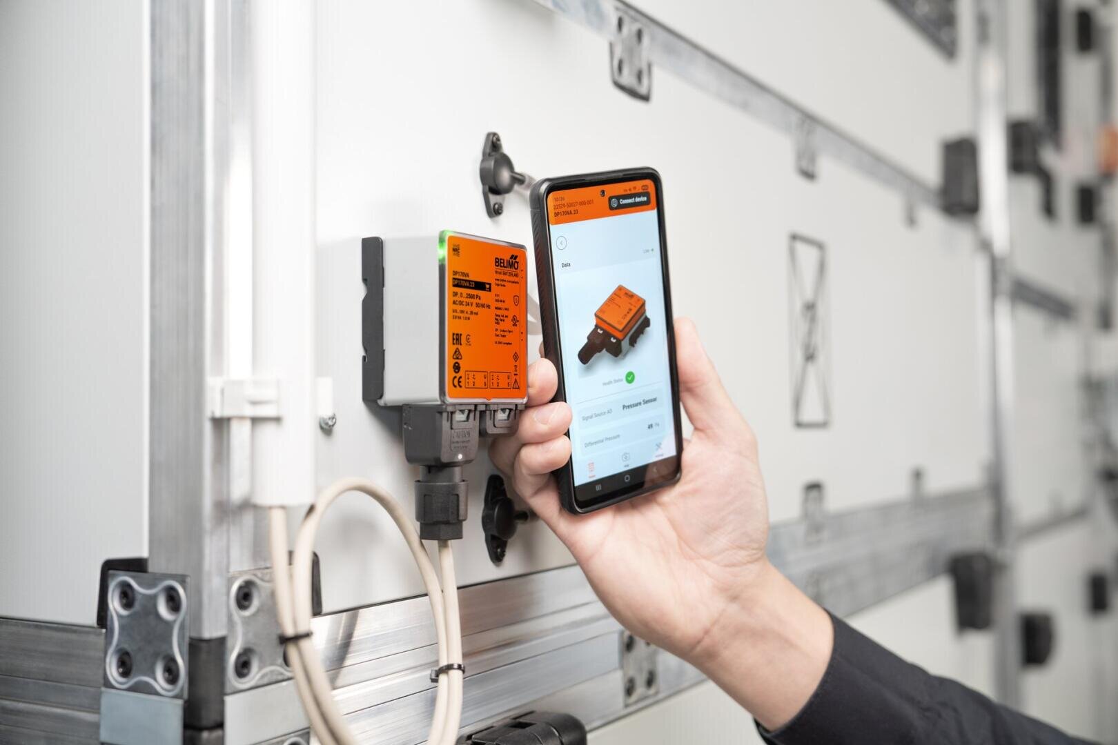 Une personne tient dans sa main un smartphone qui affiche des informations sur un appareil électronique orange monté sur un panneau industriel blanc et relié à l'appareil par des câbles.